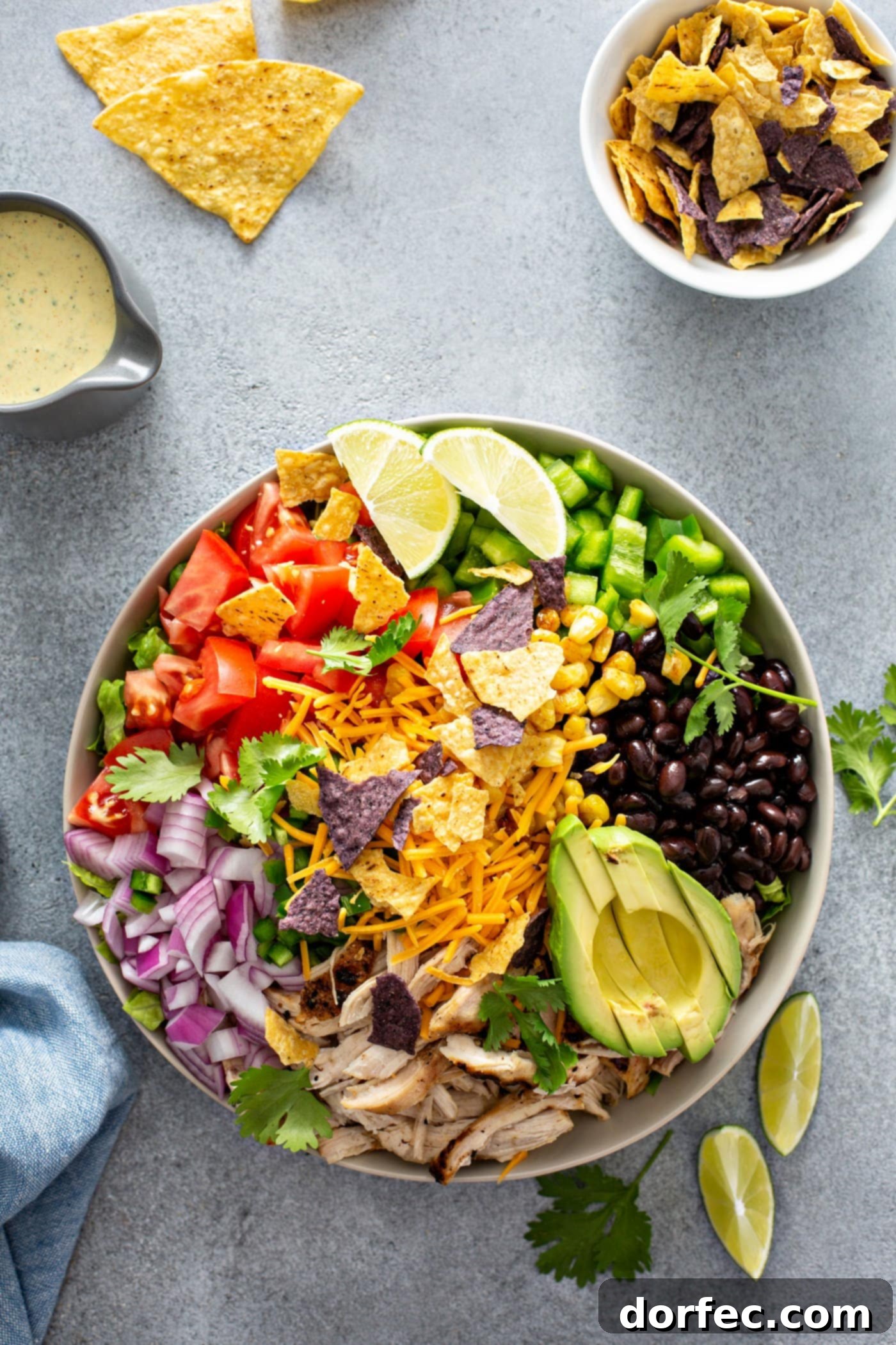 Simple Chicken Taco Salad A chicken taco salad in a large bowl. It has chopped tomatoes, red onions, cheese, cilantro, lime wedges, chicken, corn, beans and peppers. There is also a sliced avocado. There are chips and a small container of dressing in the background.