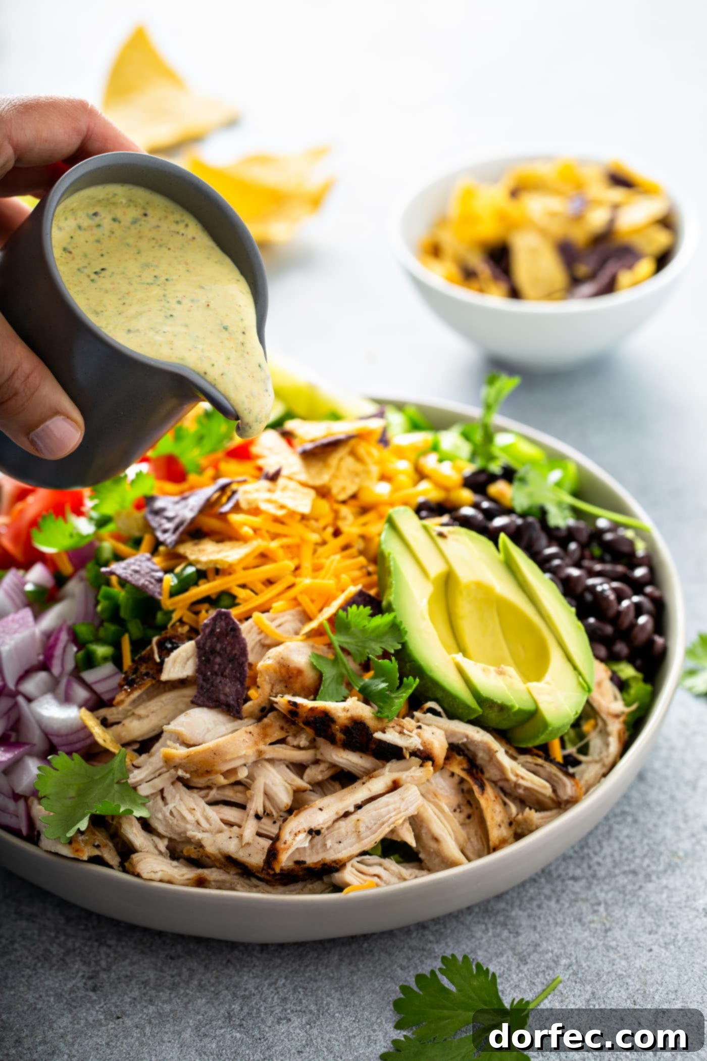 Simple Chicken Taco Salad A chicken taco salad in a large bowl. It has chopped tomatoes, red onions, cheese, cilantro, lime wedges, chicken, corn, beans and peppers. There is also a sliced avocado. There are chips in the background and dressing is being poured over the salad.