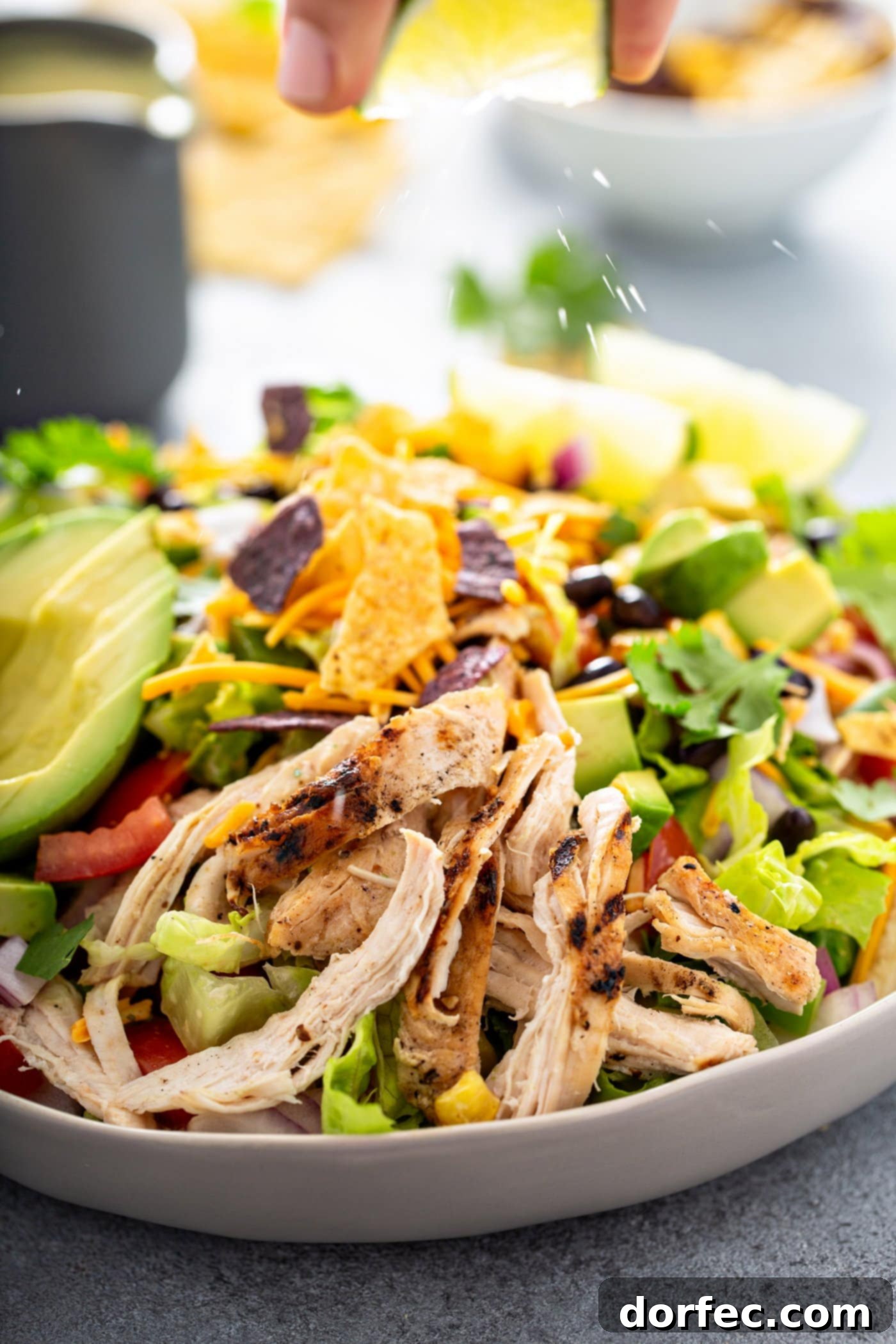 Simple Chicken Taco Salad A close-up of a large serving bowl filled with chicken taco salad. It contains shredded chicken, beans, corn, cilantro, tomatoes, red onions, and avocados. There are a couple of lime wedges next to the salad and chips and a container of dressing in the background.