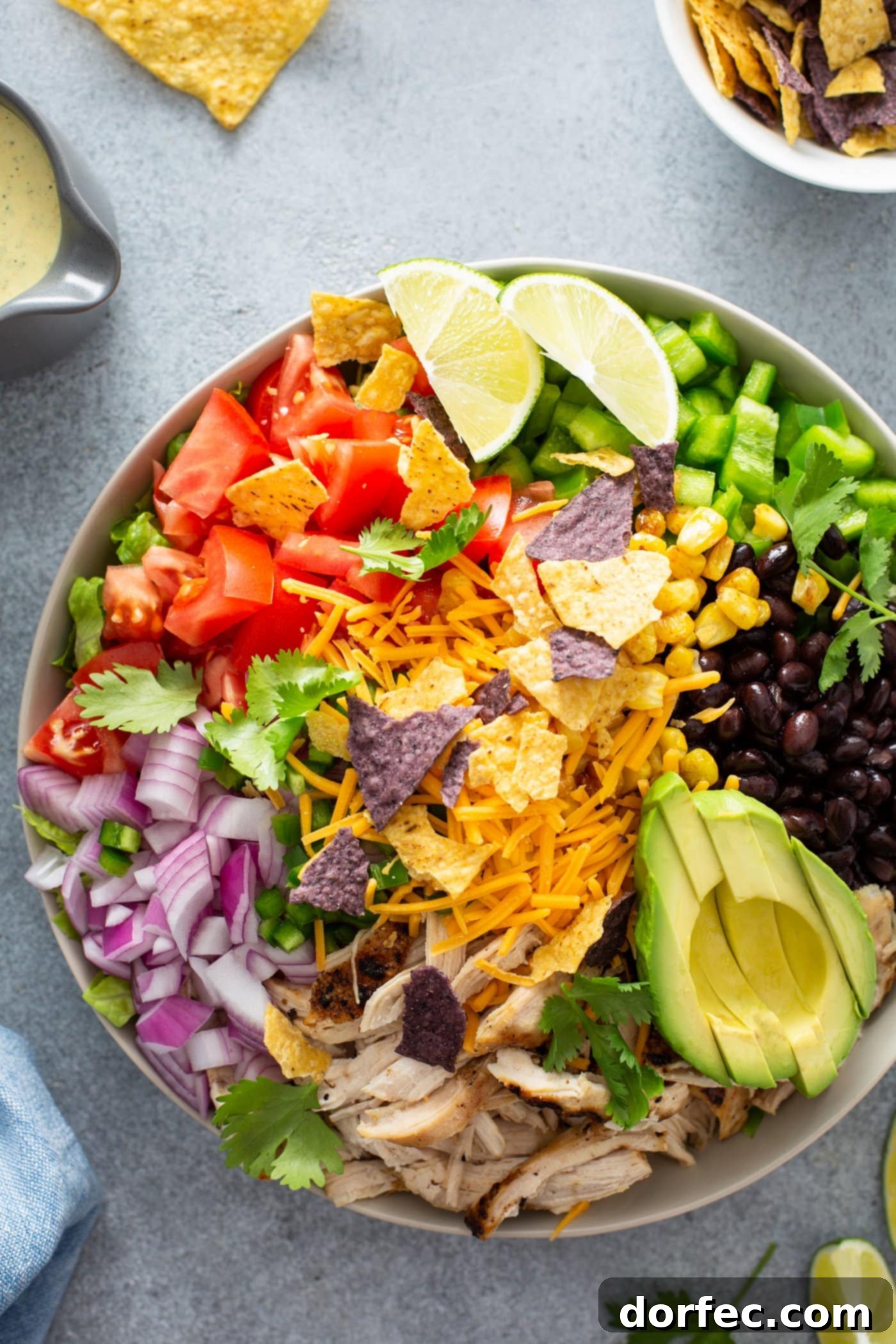 Simple Chicken Taco Salad A chicken taco salad in a large bowl. It has chopped tomatoes, red onions, cheese, cilantro, lime wedges, chicken, corn, beans and peppers. There is also a sliced avocado.