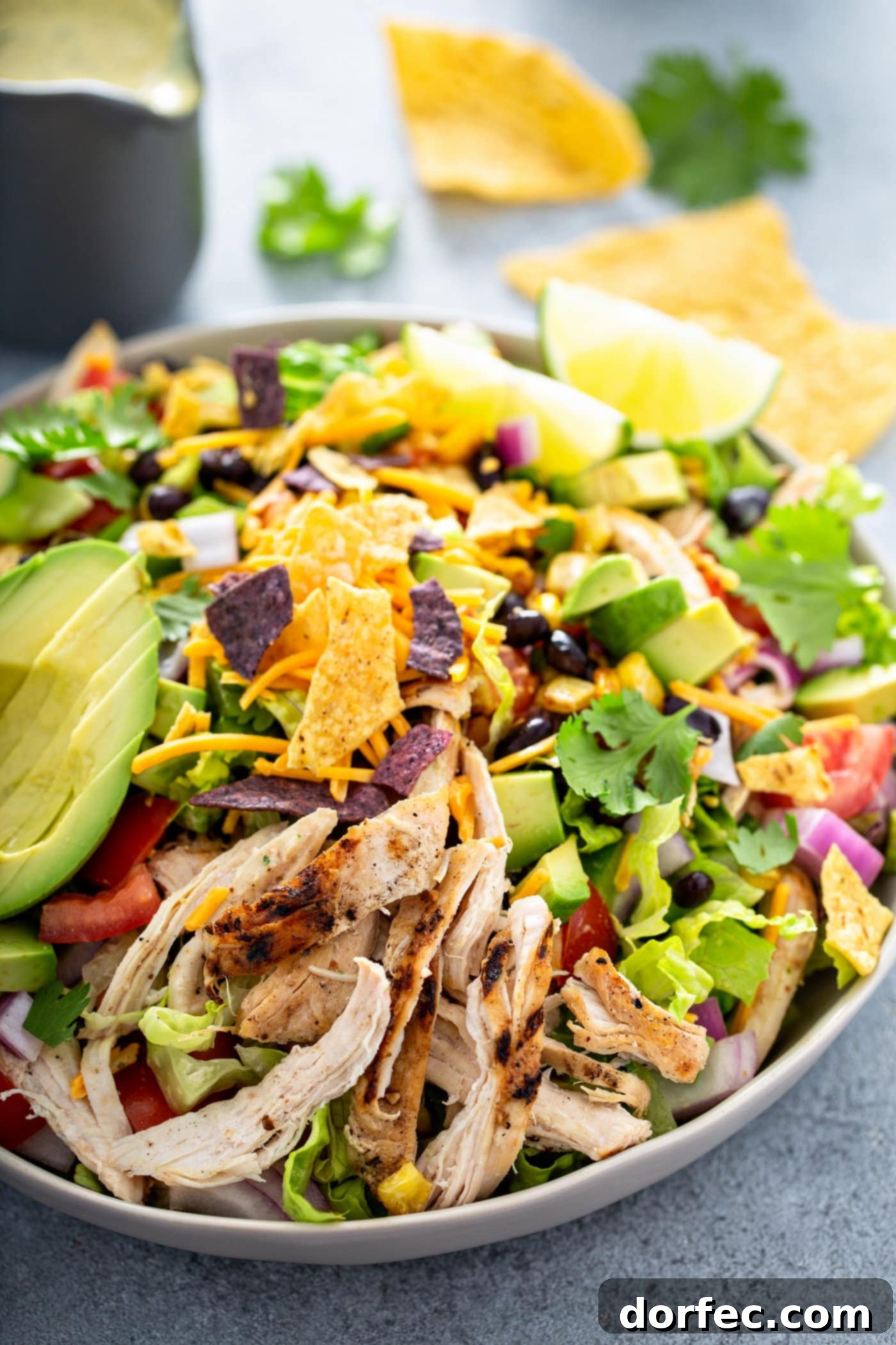 Simple Chicken Taco Salad A large bowl of chicken taco salad. There is shredded chicken, cheese, cilantro, beans, corn, tomatoes, red onions, peppers and sliced avocados. There are 2 lime wedges next to the salad and chips and dressing in the background.