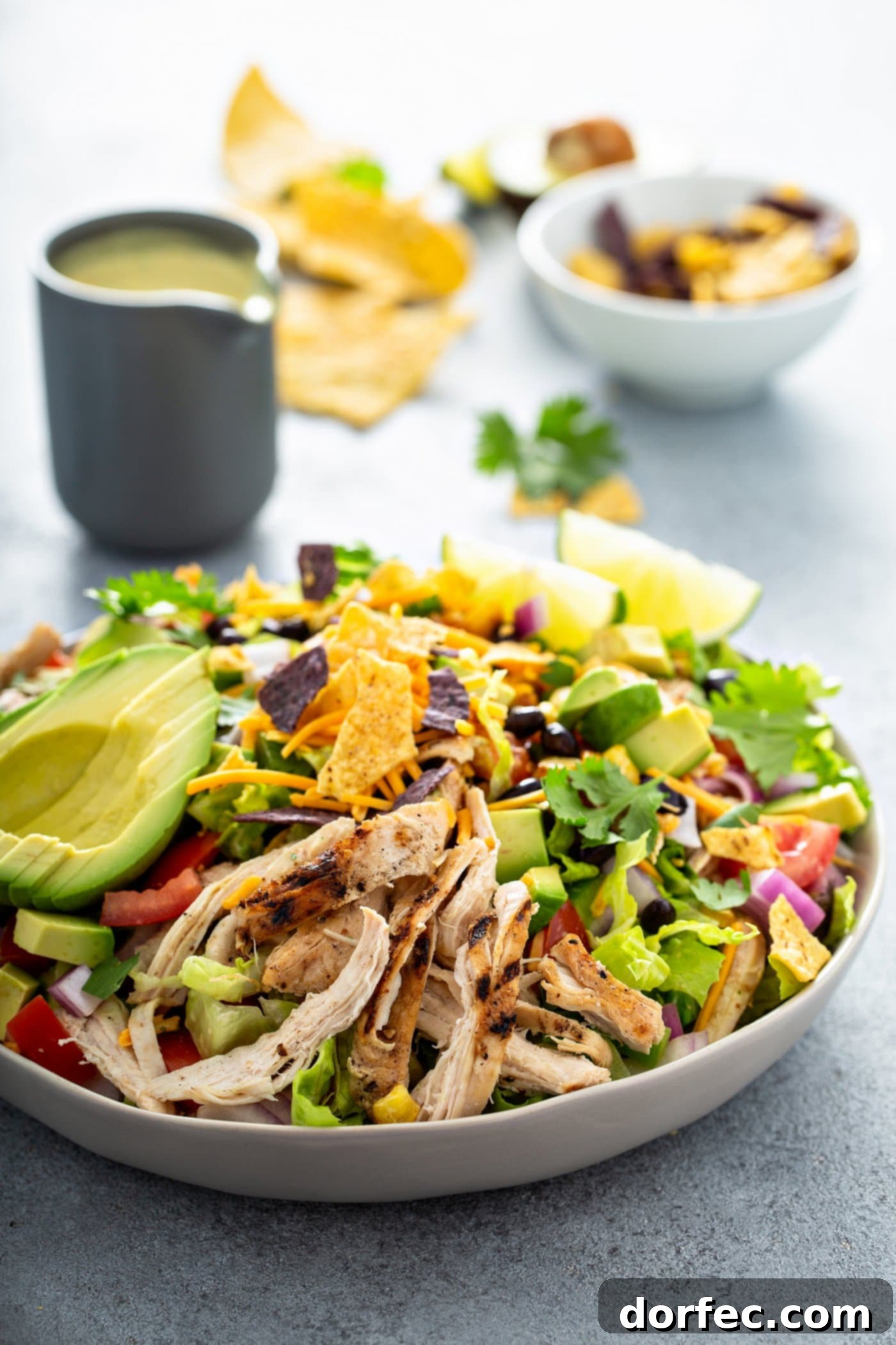 Simple Chicken Taco Salad A chicken taco salad in a large bowl. It has chopped tomatoes, red onions, cheese, cilantro, lime wedges, chicken, corn, beans and peppers. There is also a sliced avocado. There are chips and a small container of dressing in the background.