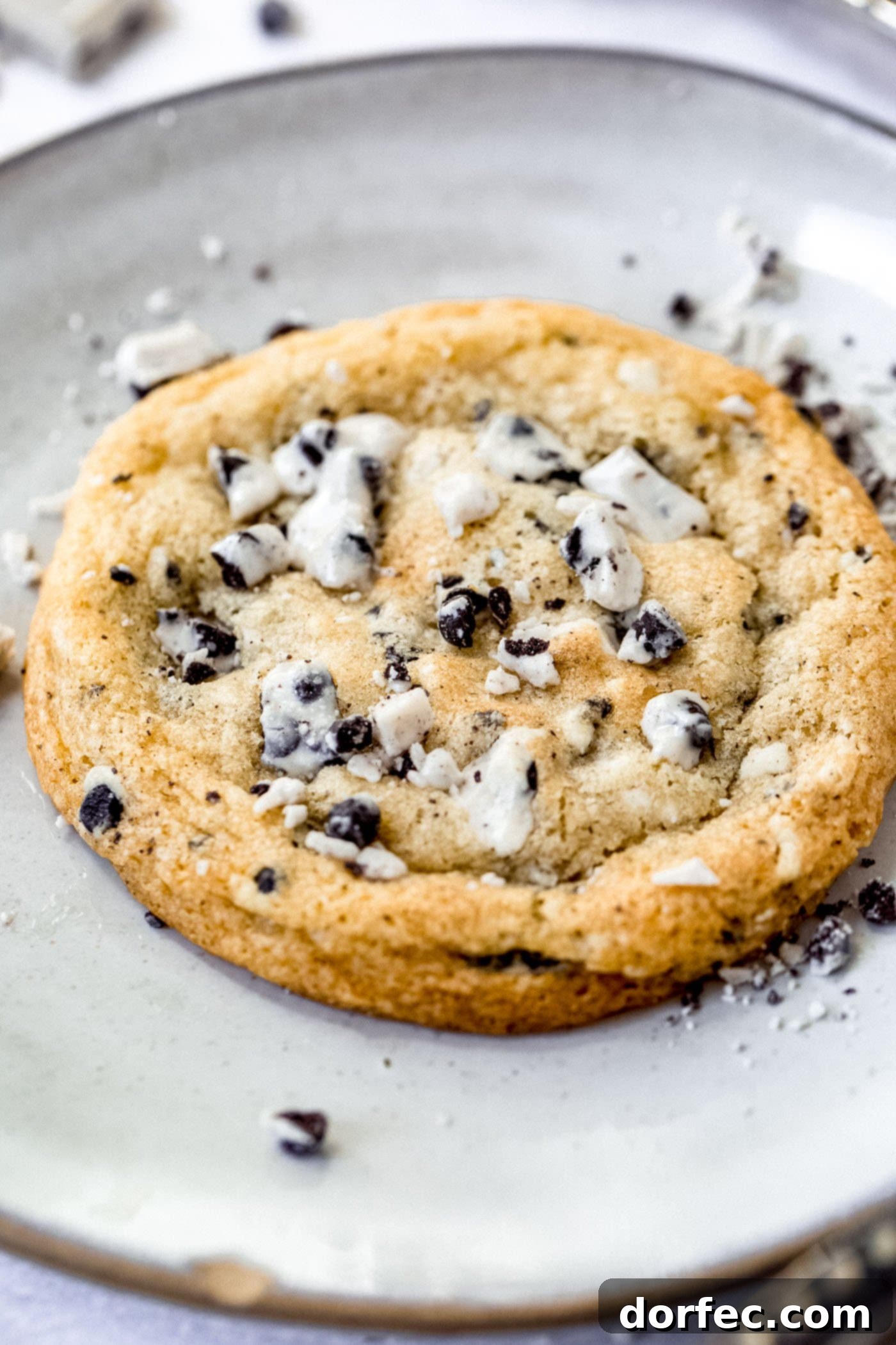 A large cookies and cream cookie on a dessert plate. There are bits of cookies and cream candy bar in the cookie and on the plate around the cookie.