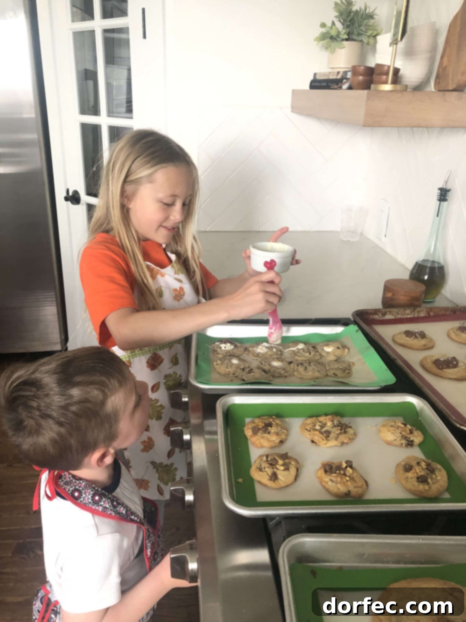 A photo of a young girl spooning melted white chocolate over chocolate chip cookies with her little brother watching.