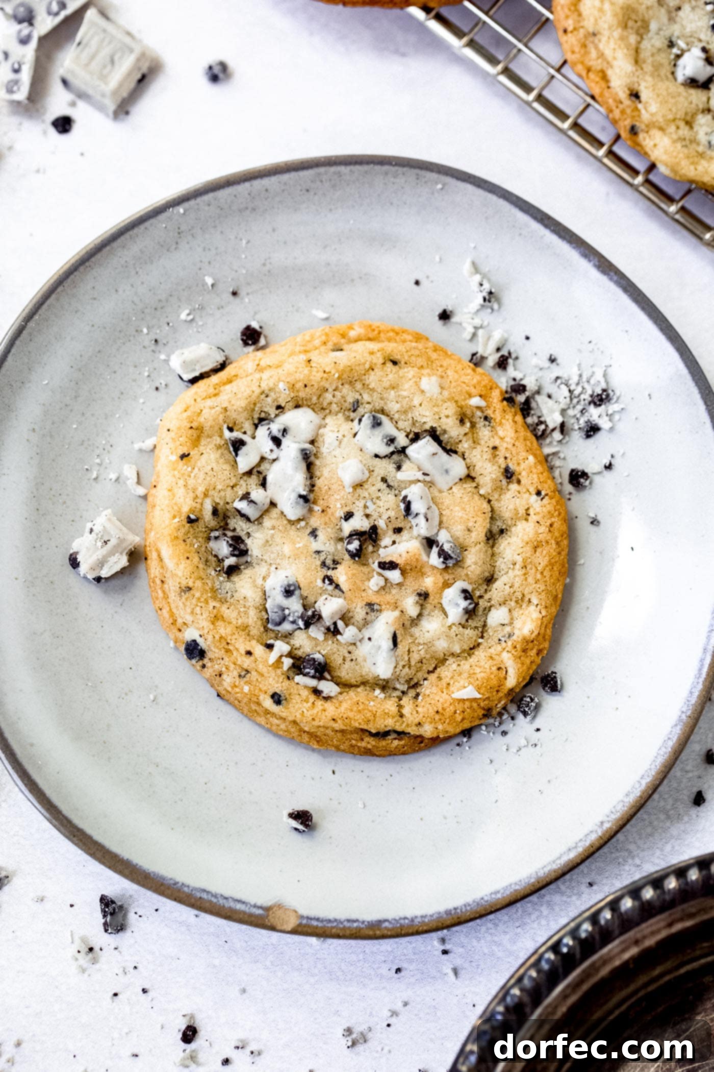 A dessert plate holding a large cookies and cream cookie. The cookie is loaded with bits of cookies and cream candy bar and there are bits of candy bar on the plate.