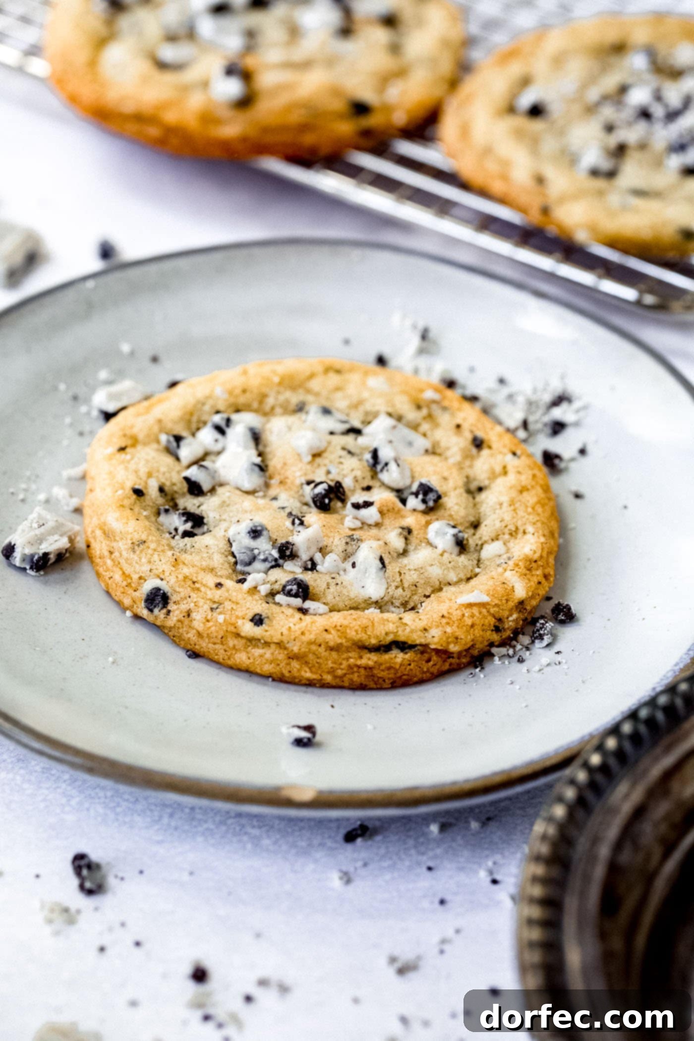 a large cookies and cream cookie on a white dessert plate. The cookie is loaded with bits of cookies and cream candy bar. there are bits of candy bar on the plate and a cooling rack of cookies in the background.