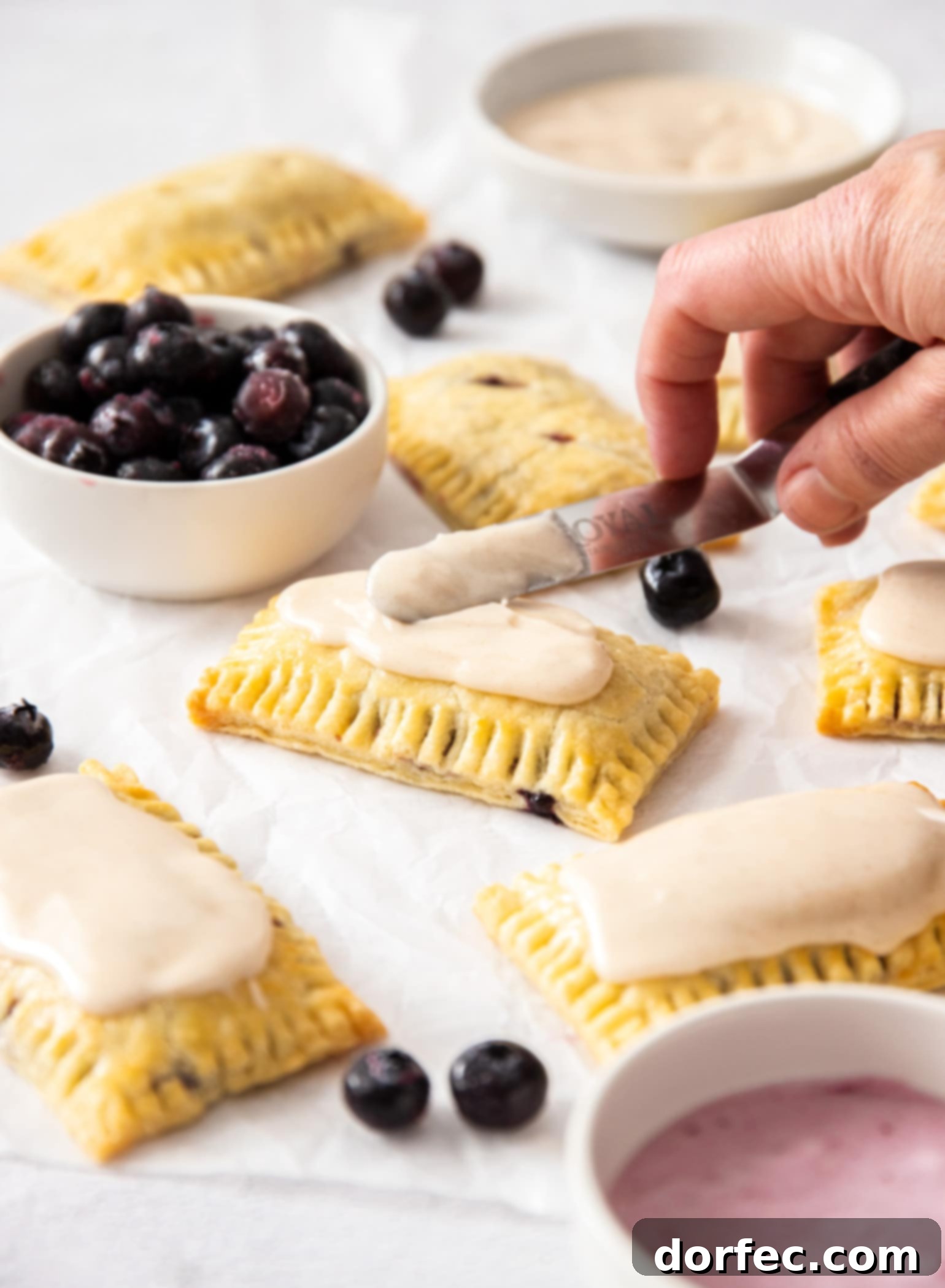 Homemade pop tarts being spread with icing. A small white bowl filled with blueberries to the side, enhancing the visual appeal.