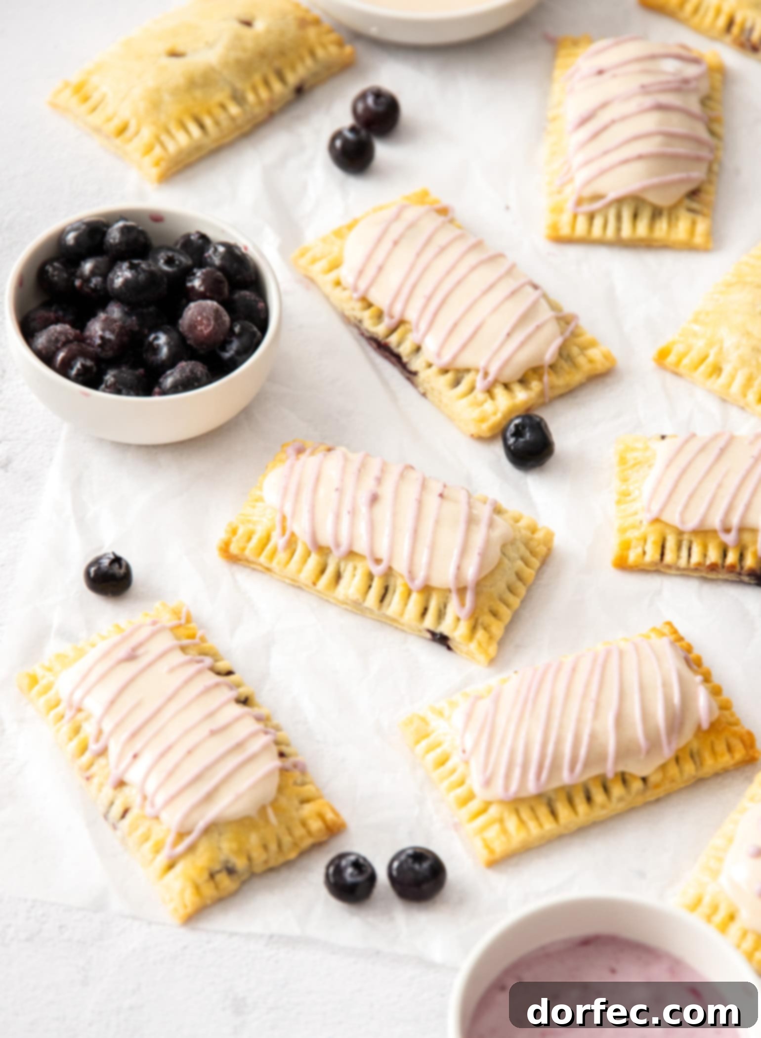 A batch of homemade pop tarts on a sheet of white baking paper. A small white bowl of blueberries, ready for a delicious breakfast.