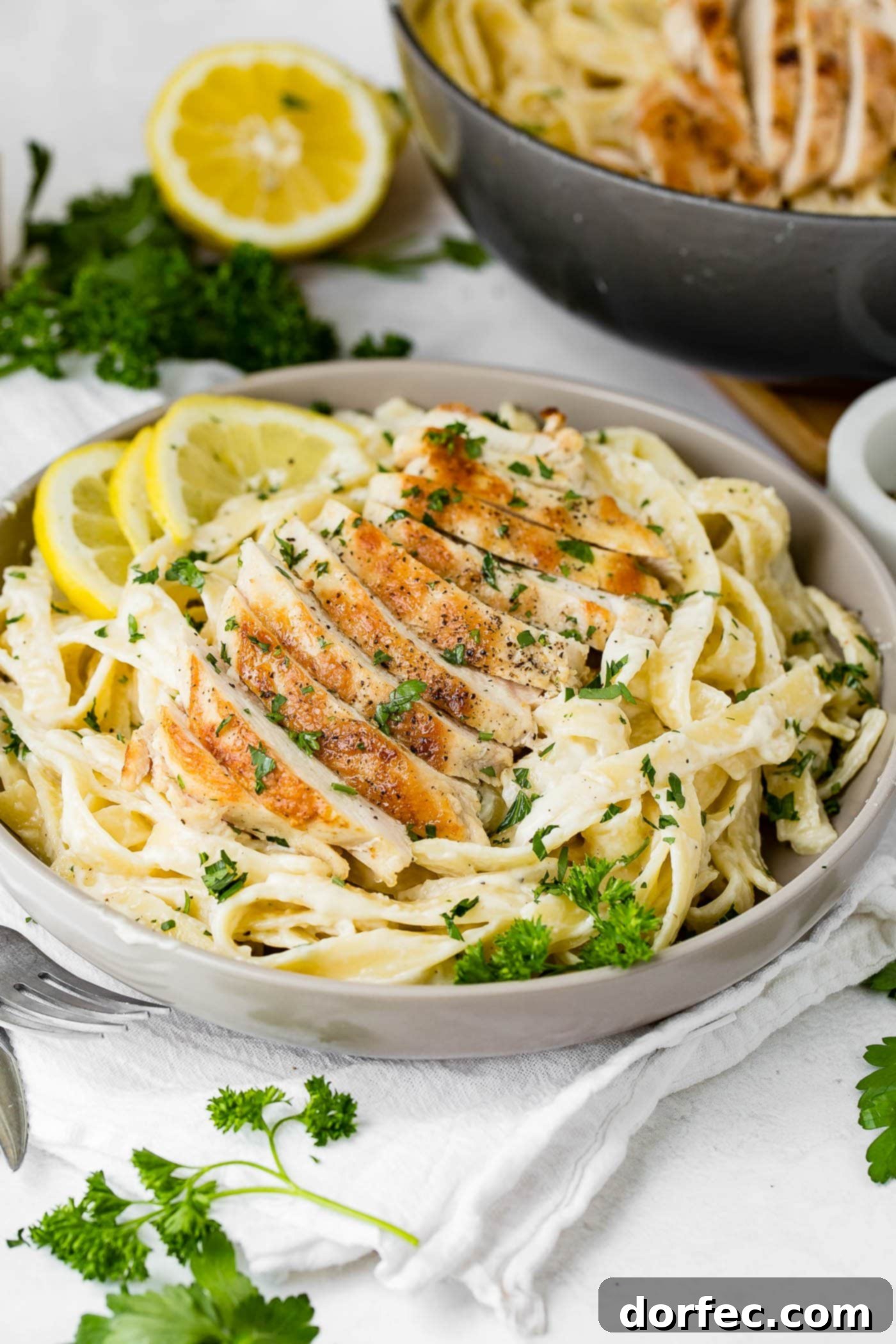 Chicken Fettuccine Alfredo A serving bowl of chicken fettaccini alfredo. There are slices of chicken on top of the pasta and it is sprinkled with fresh parsley. There are three lemon slices on the edge of the dish. Fresh parsley, lemons, and chicken are in the background.