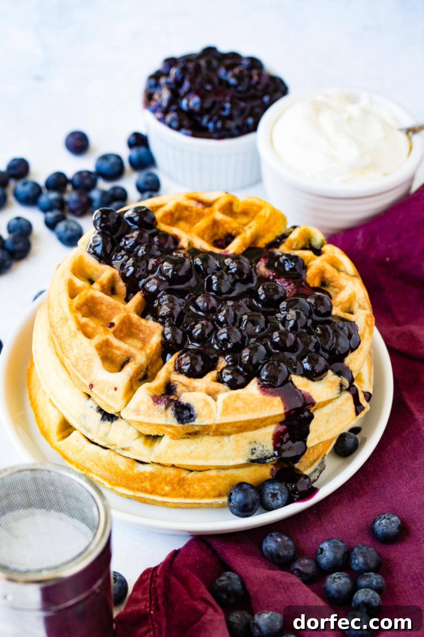 Blueberry Waffles A beautifully arranged stack of three golden blueberry waffles, generously adorned with a heap of fresh blueberries on top. In the soft-focus background, a rustic bowl of blueberries and a bowl of fluffy whipped cream complete the inviting breakfast scene, suggesting a delightful and indulgent meal.