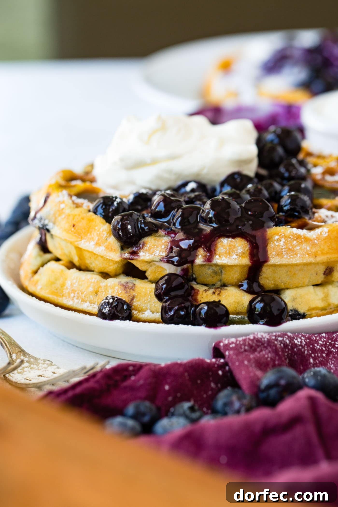 Blueberry Waffles A beautifully presented stack of fluffy blueberry waffles on a pristine white dinner plate. A generous scoop of plump, fresh blueberries cascades over the top and down the sides, complemented by a dollop of rich whipped cream. Scattered around the plate on a rustic wooden table are more fresh blueberries, creating an inviting and wholesome breakfast display.
