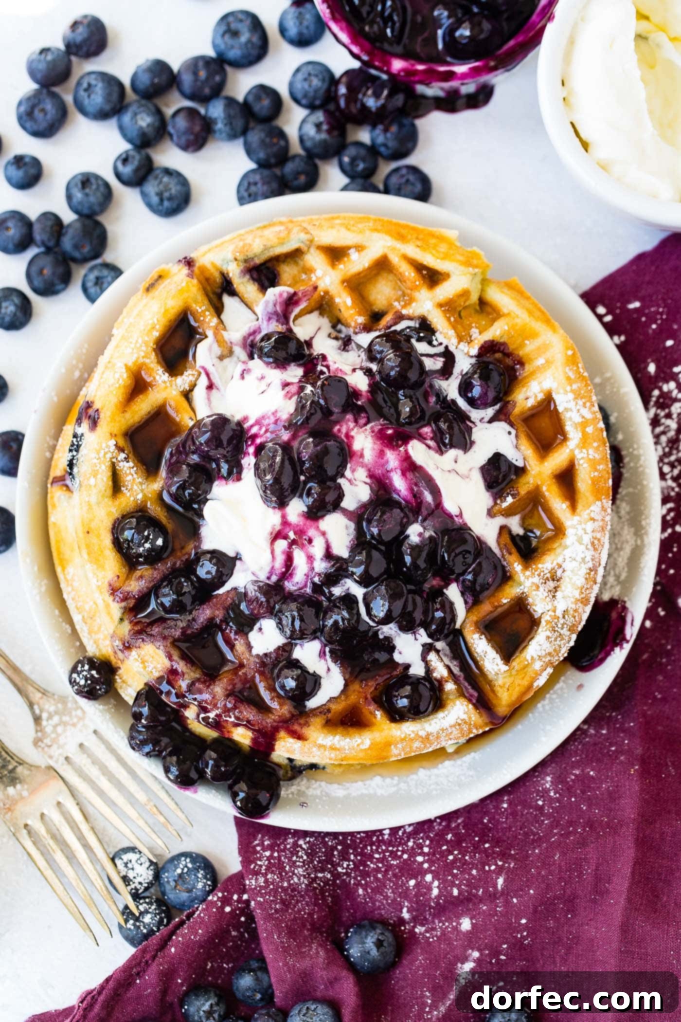 Blueberry Waffles A detailed top-down view of a single, perfectly cooked blueberry waffle, adorned with a generous dollop of whipped cream and a cluster of fresh blueberries. The setting includes a small white bowl filled with fresh blueberries, another bowl containing mashed blueberries with a spoon, and a third bowl of whipped cream, all arranged on a wine-colored napkin beside two elegant forks, creating an inviting tableau of breakfast indulgence.