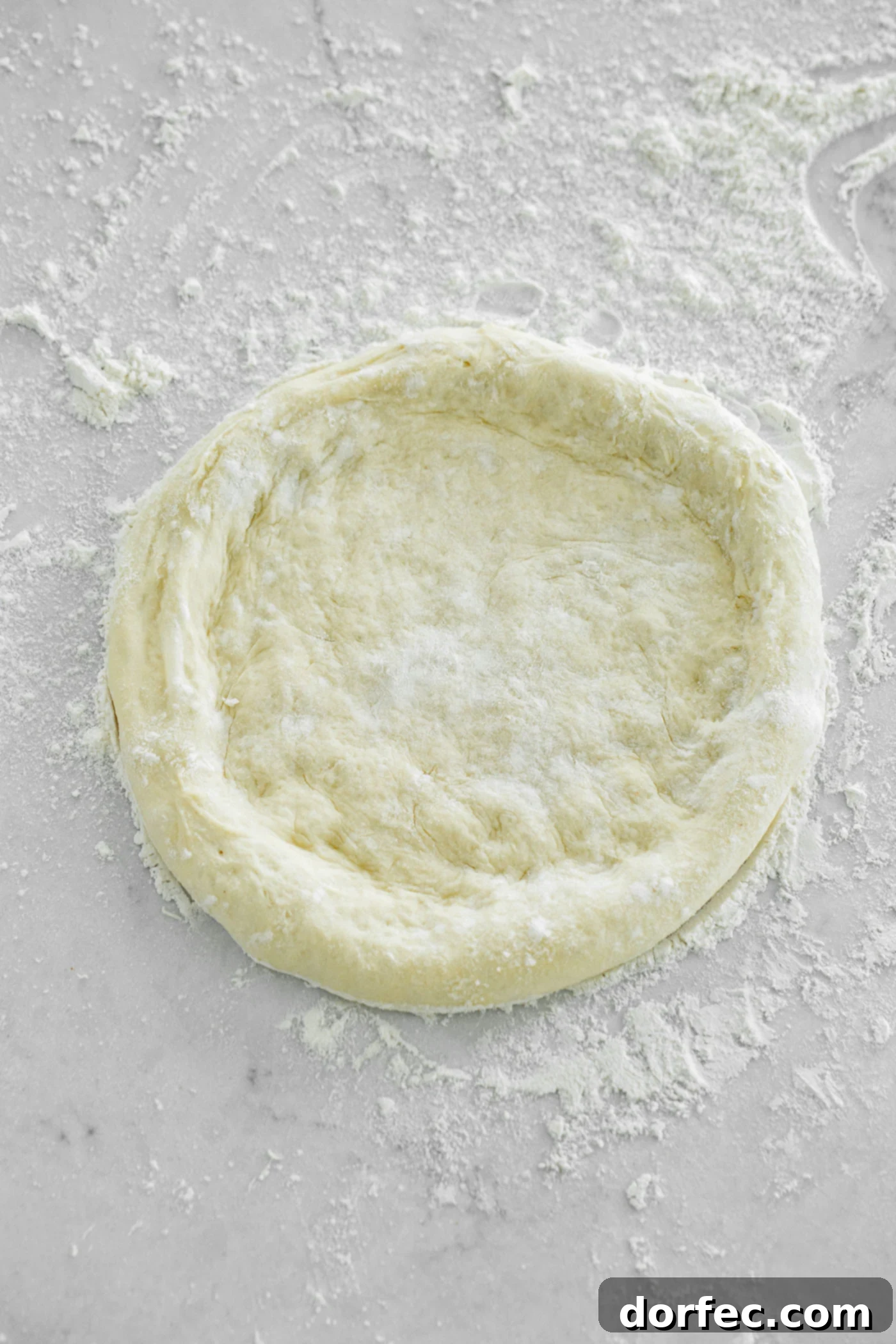Homemade pizza dough that has been formed into a round pizza shape. the edges are rounded like a thick crust pizza. The dough is on a floured surface.