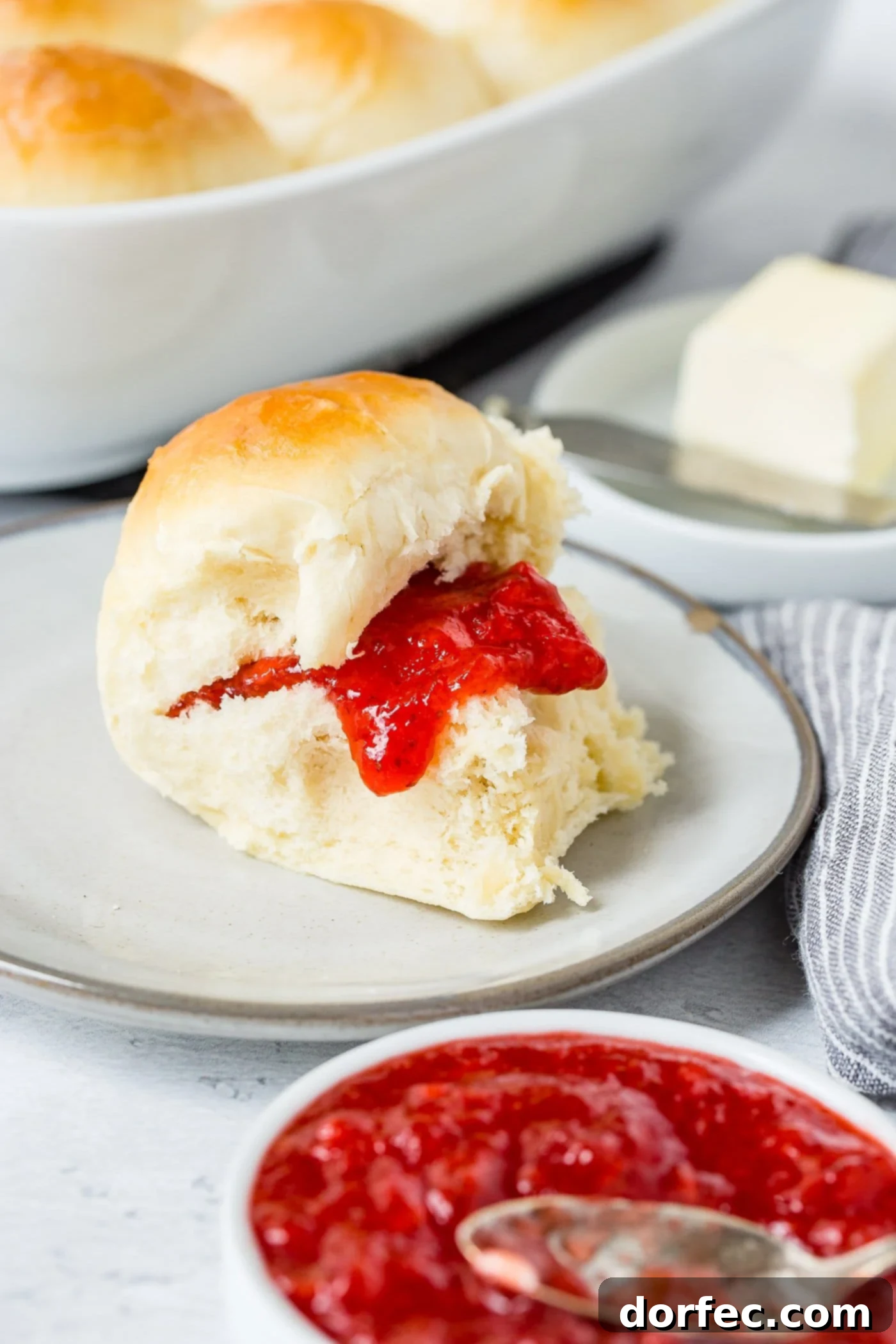 A white dinner roll on a small plate. There is strawberry jam on the roll. A dish of butter and a dish of jam are next to the roll.