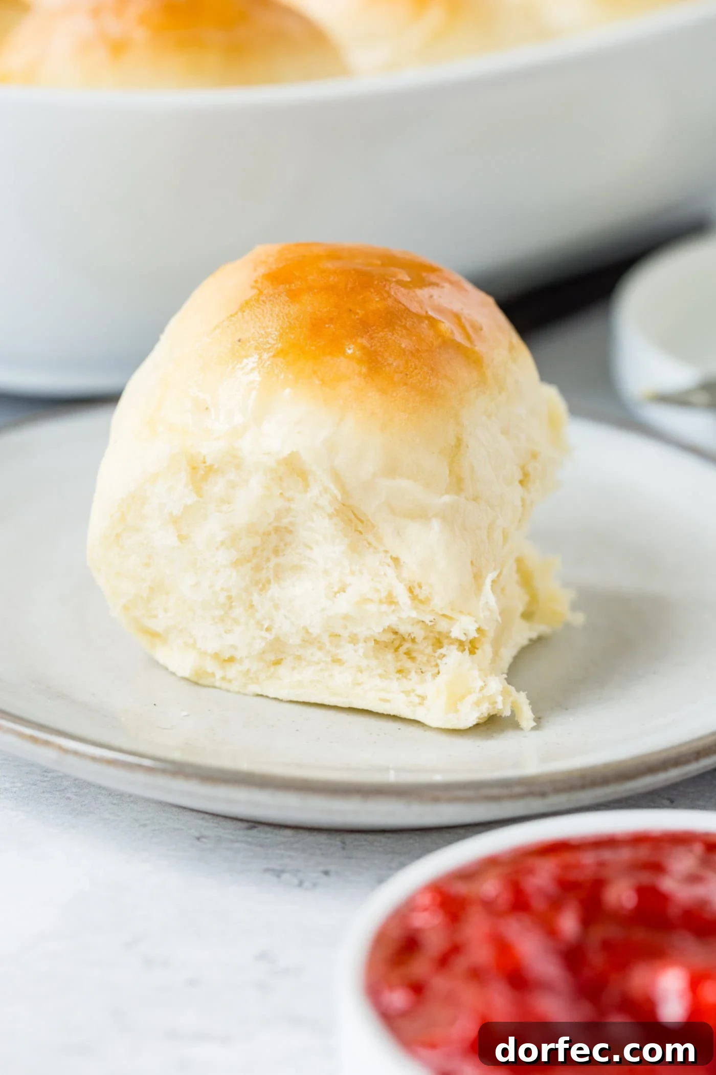 A white dinner roll on a small plate. The roll is perfectly baked to a golden brown. A small dish of strawberry jam is next to the roll.