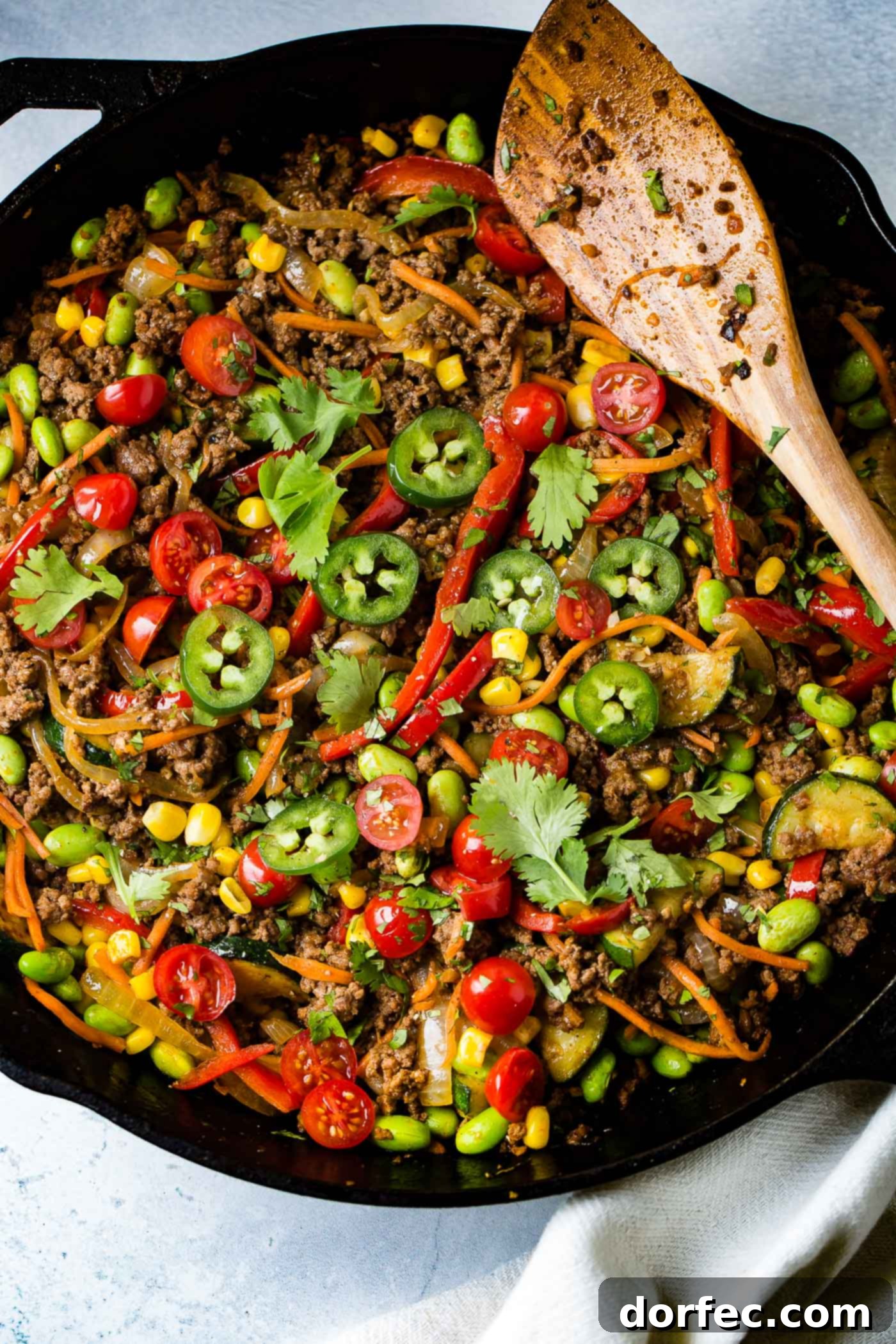 Taco skillet. There is a wooden spoon laying across the cast iron skillet. There are sliced tomatoes, sliced jalapeno, carrots, corn, pasta and ground beef.