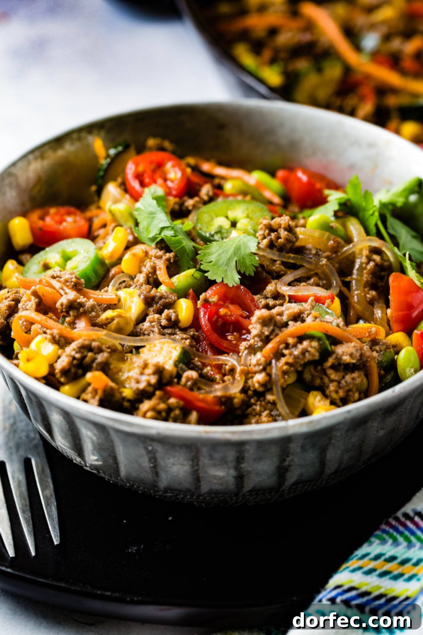 A bowl of taco pasta skillet. There are sliced tomatoes, sliced jalapenos, ground beef, zucchini, carrots, and fresh parsley.