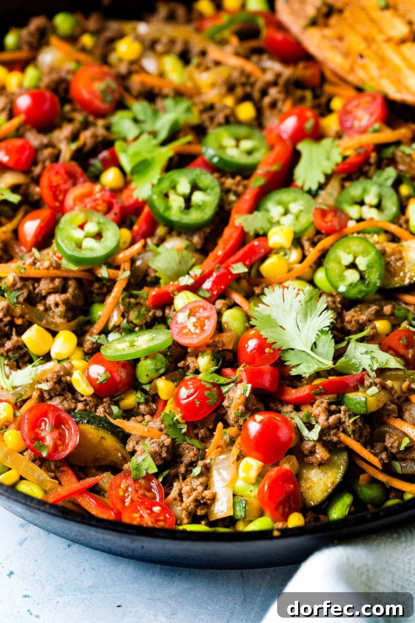 A close up of a taco skillet. It is full of sliced tomatoes, sliced jalapeno, carrots, zucchini, corn and ground beef.