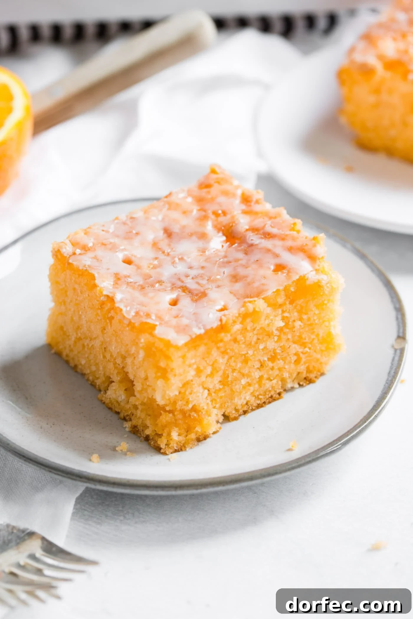 A photo of a piece of orange jello cake on a small white plate with another serving in the background and a fork in the foreground, highlighting its moist texture and bright glaze.