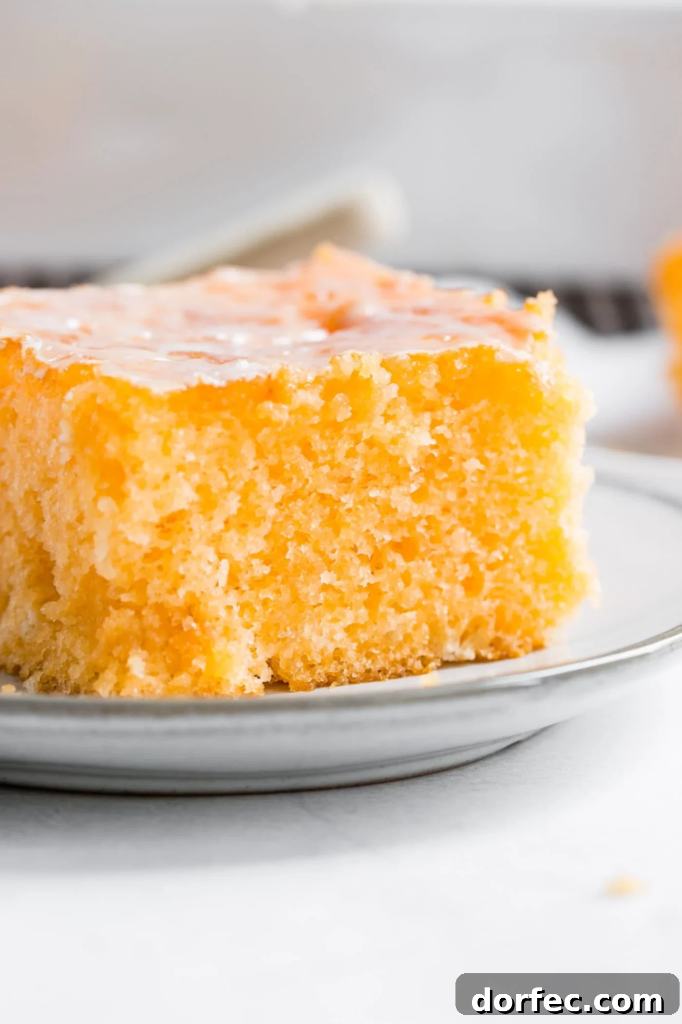 A closeup photo of a piece of orange jello cake on a small white plate, showing the texture of the cake and the glossy glaze.