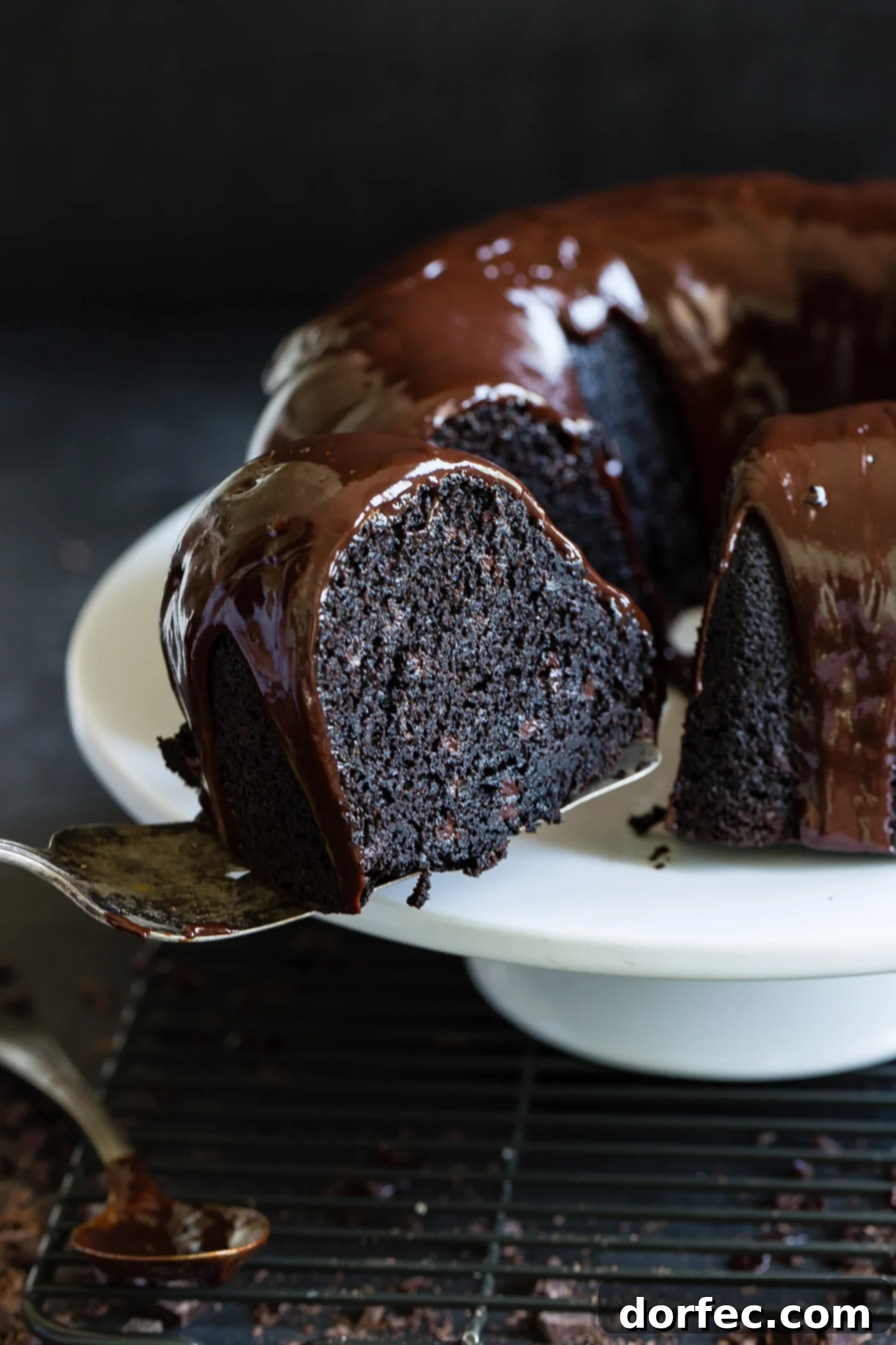 A slice of zucchini chocolate bundt cake being removed from the cake. The inside of the cake is dark, moist chocolate and there is a chocolate glaze on top of the cake.