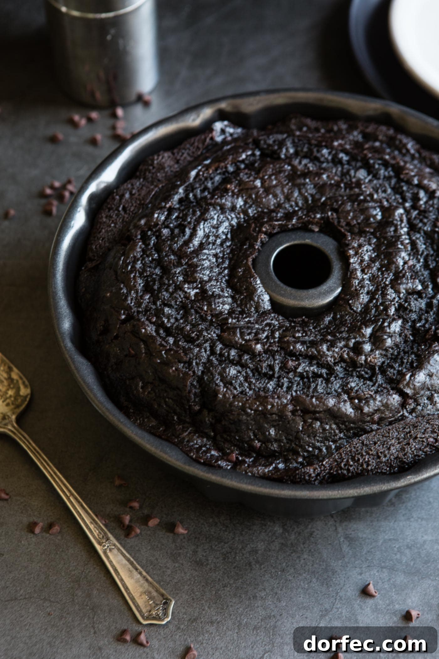 A baked zucchini chocolate bundt cake in the bundt pan. Bits of chocolate and a cake server are next to the cake. 