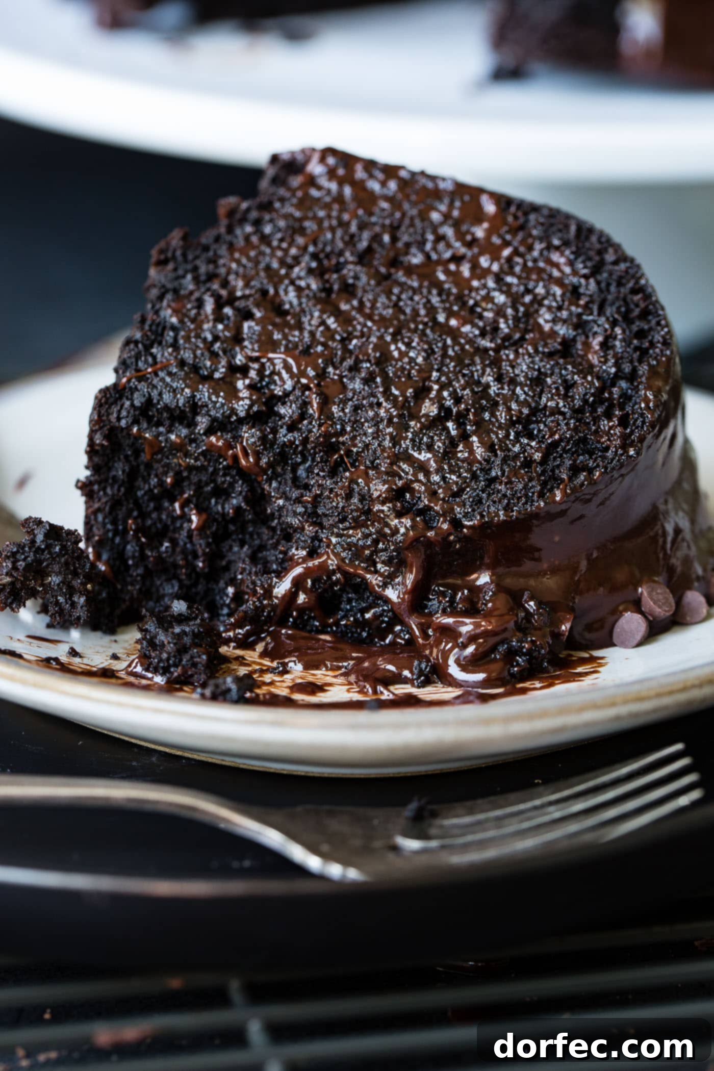 A slice of zucchini chocolate bundt cake on a white dessert plate. The cake is dark, moist chocolate with a chocolate glaze. A fork is next to the plate.