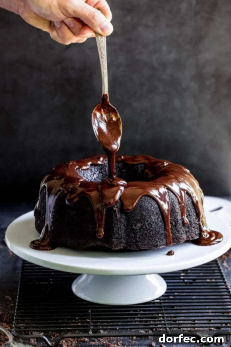A spoon with thick cocoa glaze drizzling over the top of a cake which is on a white cake plate.