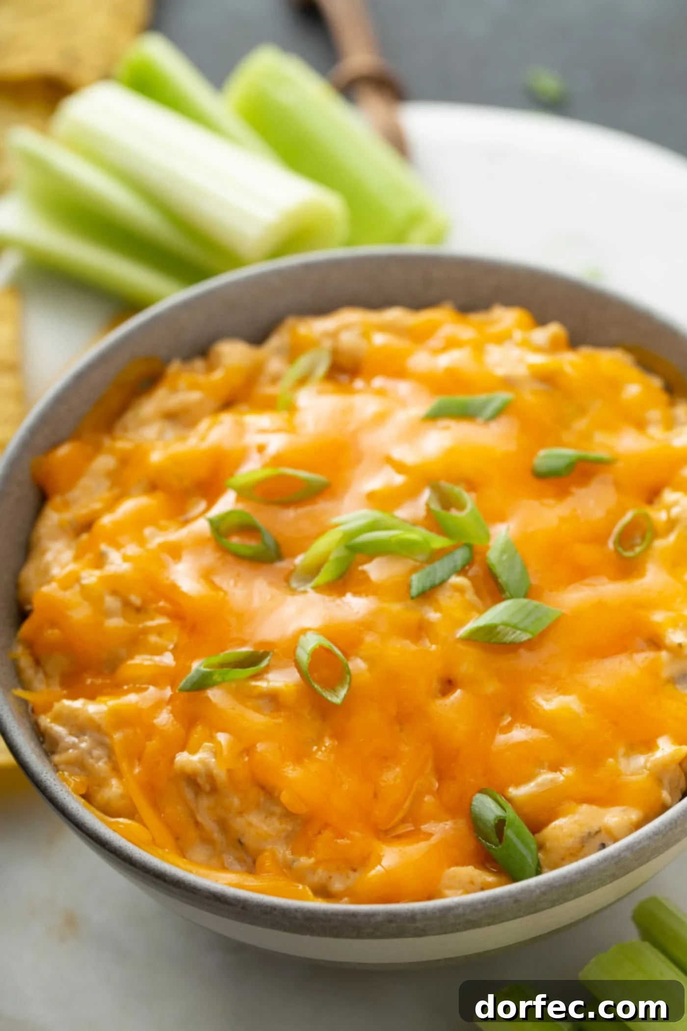 Hot Buffalo Chicken Dip with Celery A festive bowl of warm, bubbling buffalo chicken dip, crowned with melted cheese and a sprinkle of chopped green onions. Crisp celery sticks are arranged invitingly in the background, ready to be dipped.