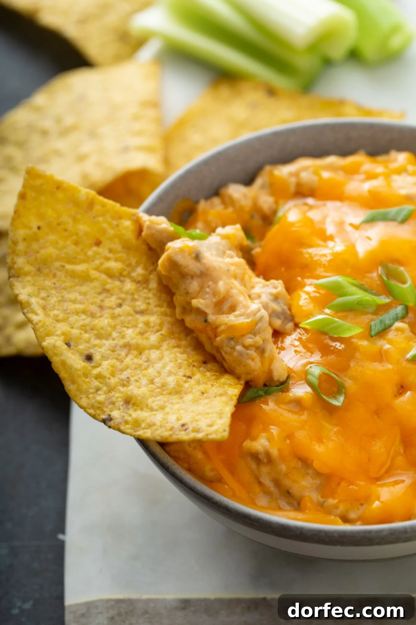 Dipping into Buffalo Chicken Dip A hand dipping a chip into warm buffalo chicken dip, highlighting the melted cheese and a sprinkle of chopped green onions on top. The moment of pure dipping satisfaction.
