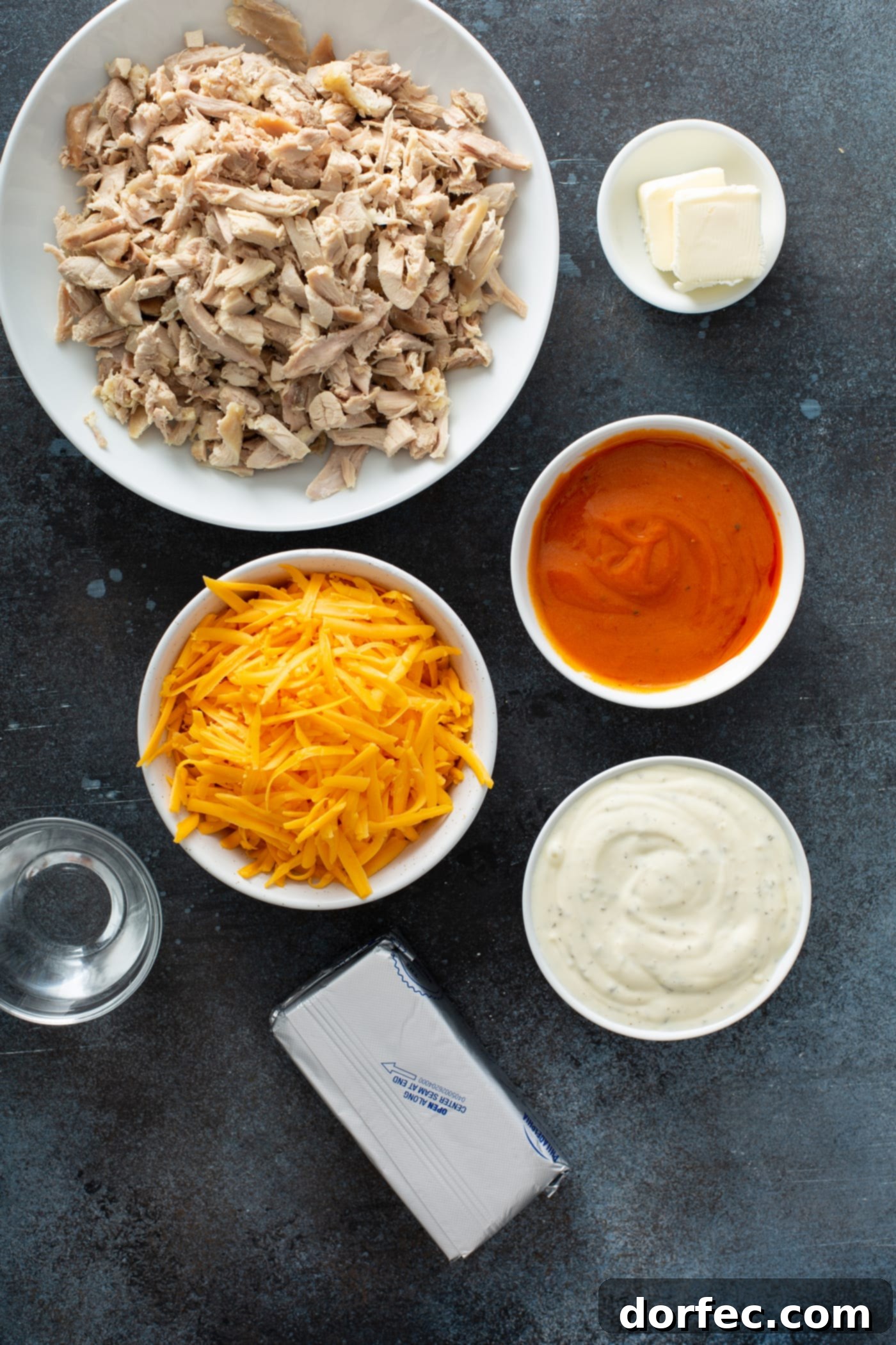 Ingredients Ready for Slow Cooker Buffalo Chicken Dip Small containers holding the various ingredients for buffalo chicken dip, including shredded cheddar cheese, cooked and shredded chicken, softened cream cheese, fiery buffalo sauce, creamy ranch dressing, and a measured amount of water.