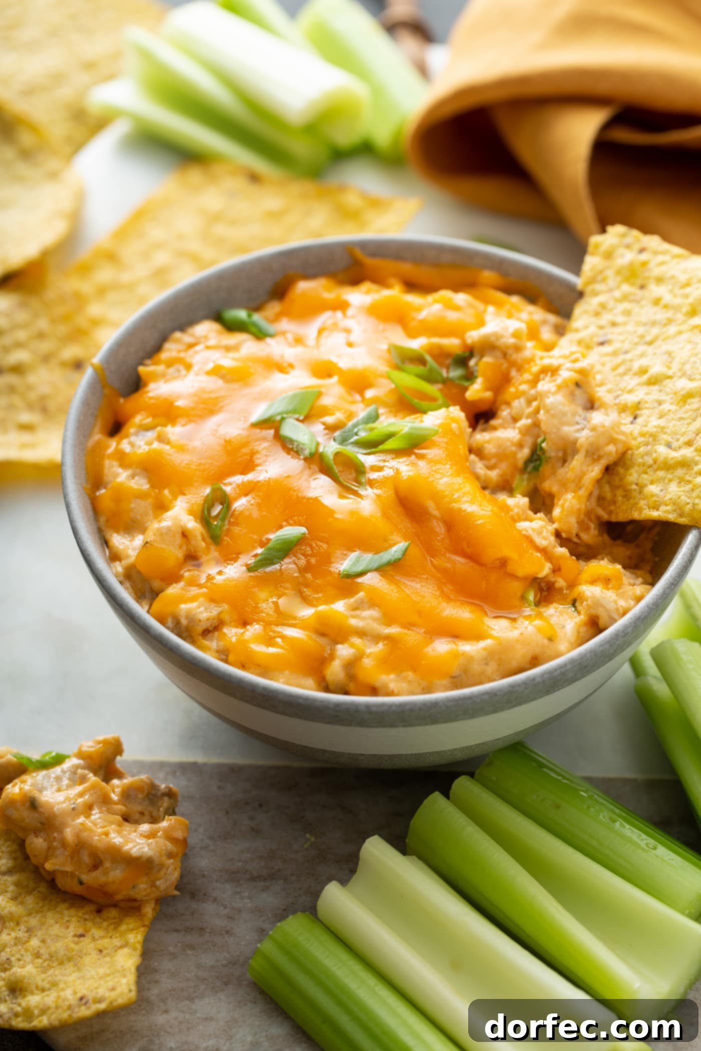Serving Slow Cooker Buffalo Chicken Dip with Dippers A beautifully presented bowl of buffalo chicken dip, generously topped with melted cheese and fresh chopped green onions. A chip is poised mid-dip, surrounded by an inviting arrangement of tortilla chips and crisp celery sticks, highlighting the perfect serving presentation.
