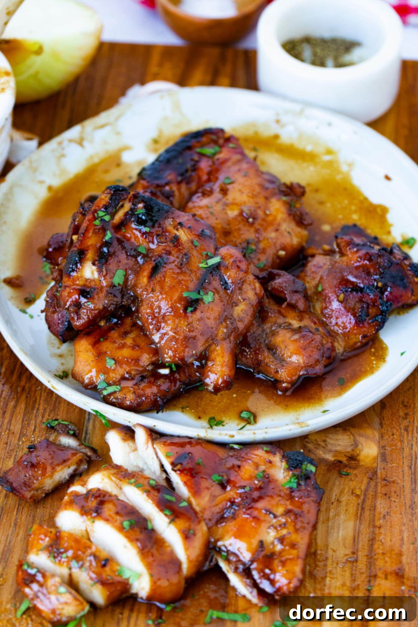 A serving plate filled with juicy marinated smoked chicken thighs. Copped parsley is sprinkled on top of the chicken and a sliced chicken thigh is on the cutting board next to the plate of chicken.