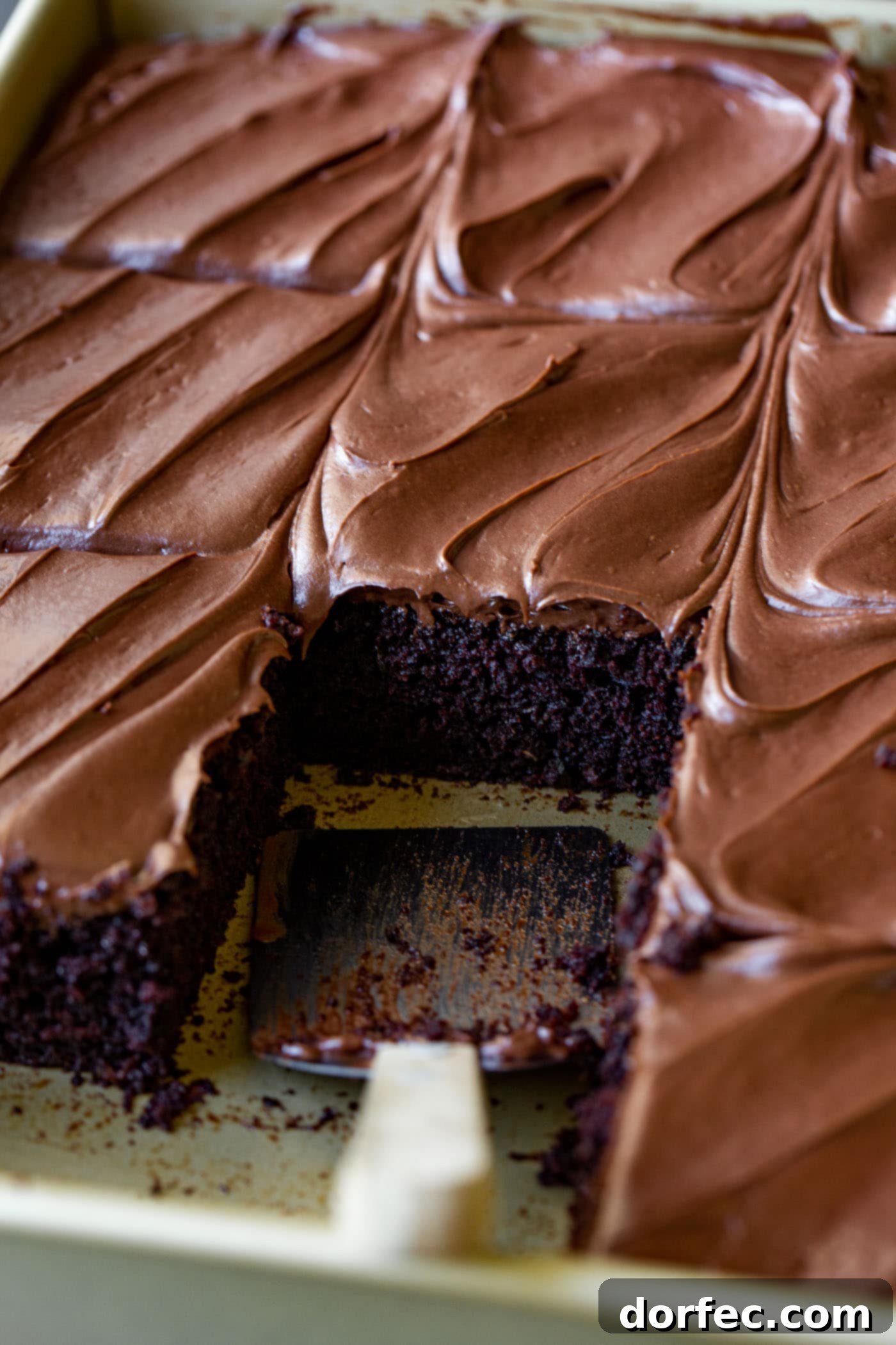 A baking pan filled with freshly baked chocolate zucchini cake, expertly cut into squares. One piece has been removed, revealing the dark, moist crumb and a generous layer of creamy chocolate frosting.