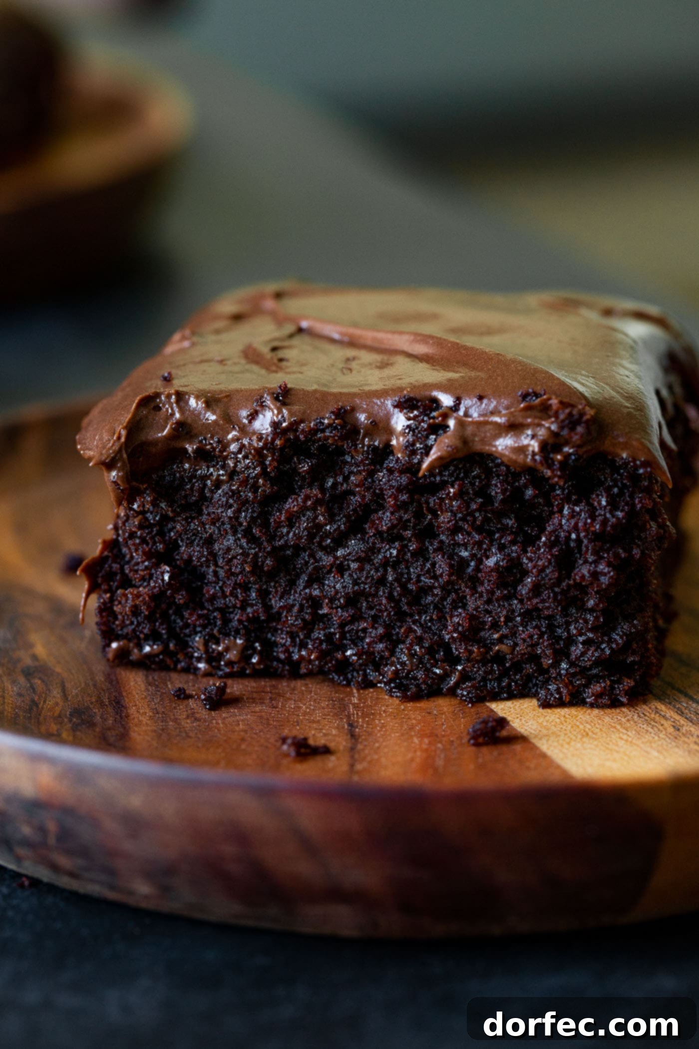 A single slice of moist chocolate zucchini cake, perfectly plated on a small wooden dessert plate. The cake's dark, rich crumb is visible, topped with a thick, creamy layer of chocolate cream cheese frosting, inviting a delightful bite.