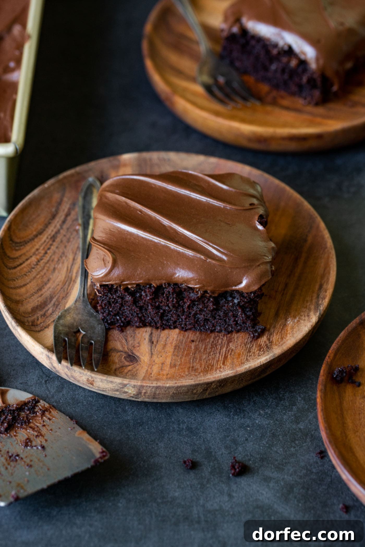 A delectable slice of moist chocolate zucchini cake presented on a wooden plate, accompanied by a fork. In the background, other plates with cake hint at a gathering, highlighting the cake's rich, dark, and inviting texture.
