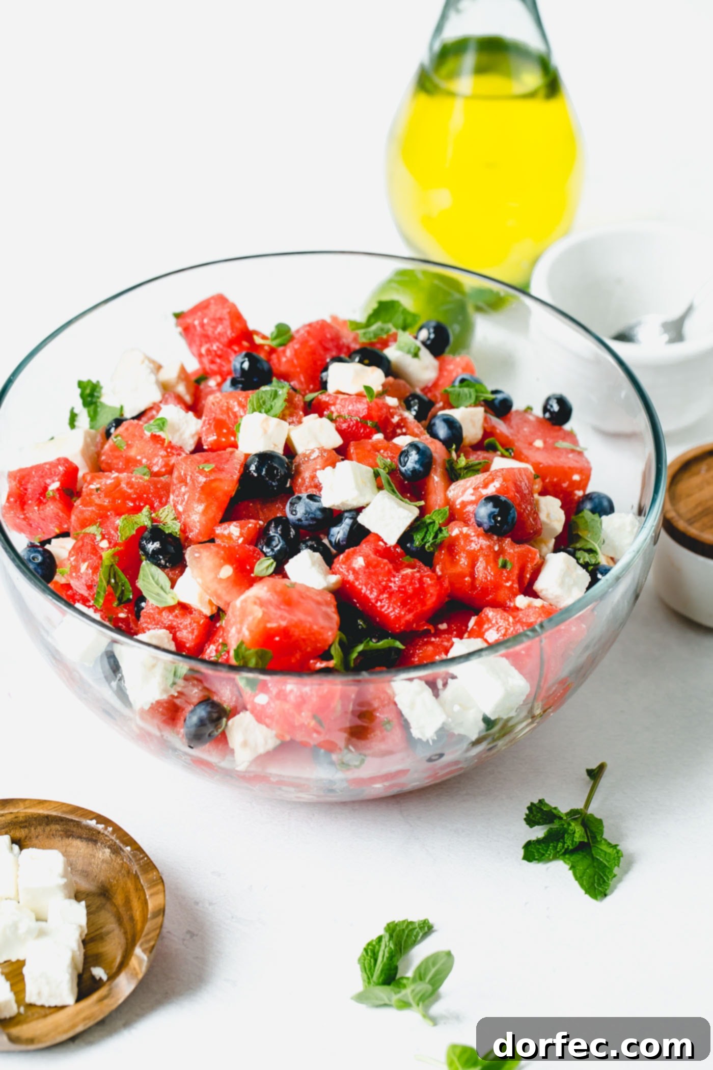 Blueberry Feta Watermelon Salad A glass bowl of blueberry feta watermelon salad featuring vibrant red watermelon chunks, plump blueberries, and creamy feta cheese cubes, garnished with fresh basil and mint leaves.