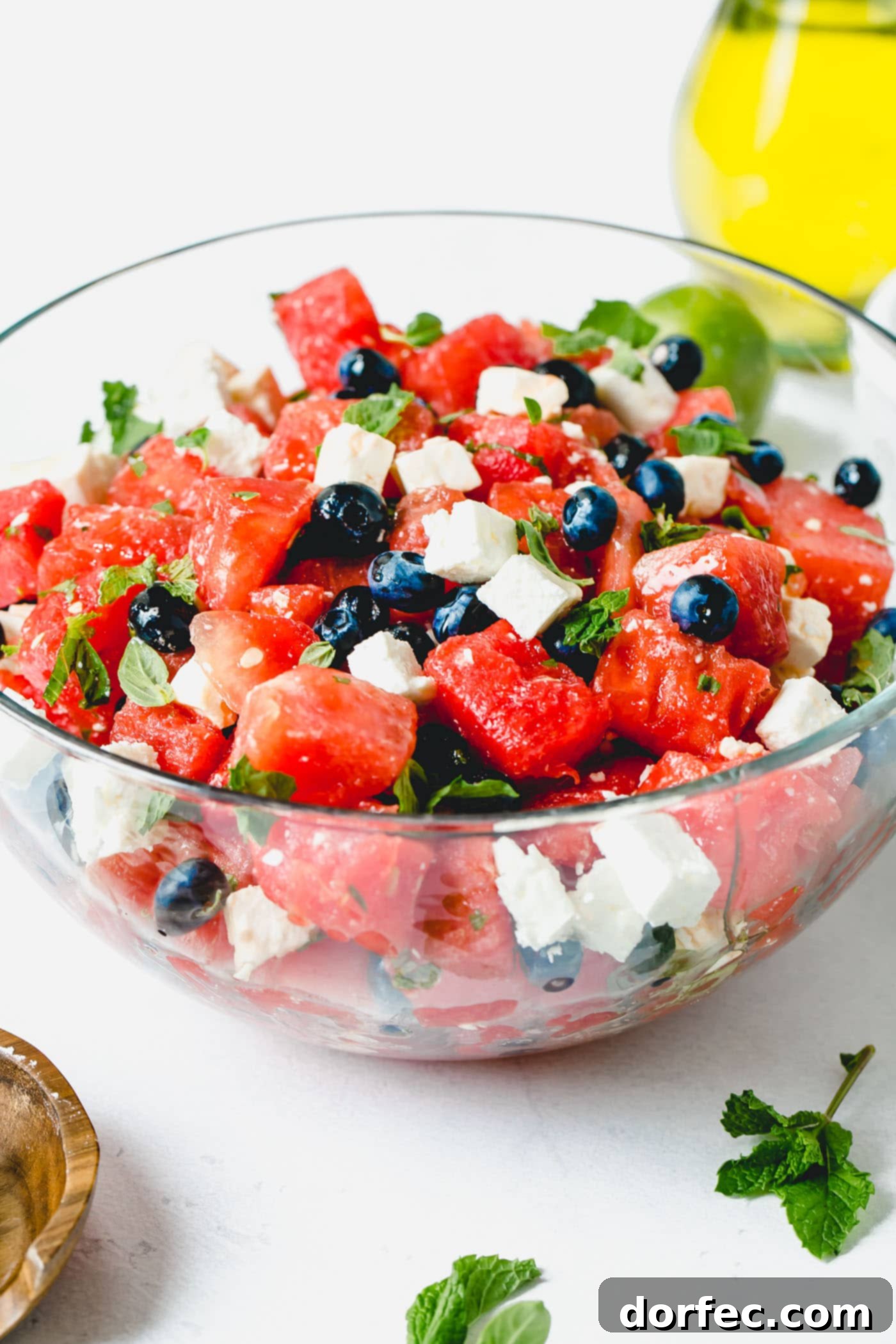 Blueberry Feta Watermelon Salad A beautiful glass serving bowl showcasing the delightful Blueberry Feta Watermelon Salad, filled with juicy watermelon cubes, vibrant blueberries, and creamy feta cheese, all mixed with fresh basil and mint leaves.