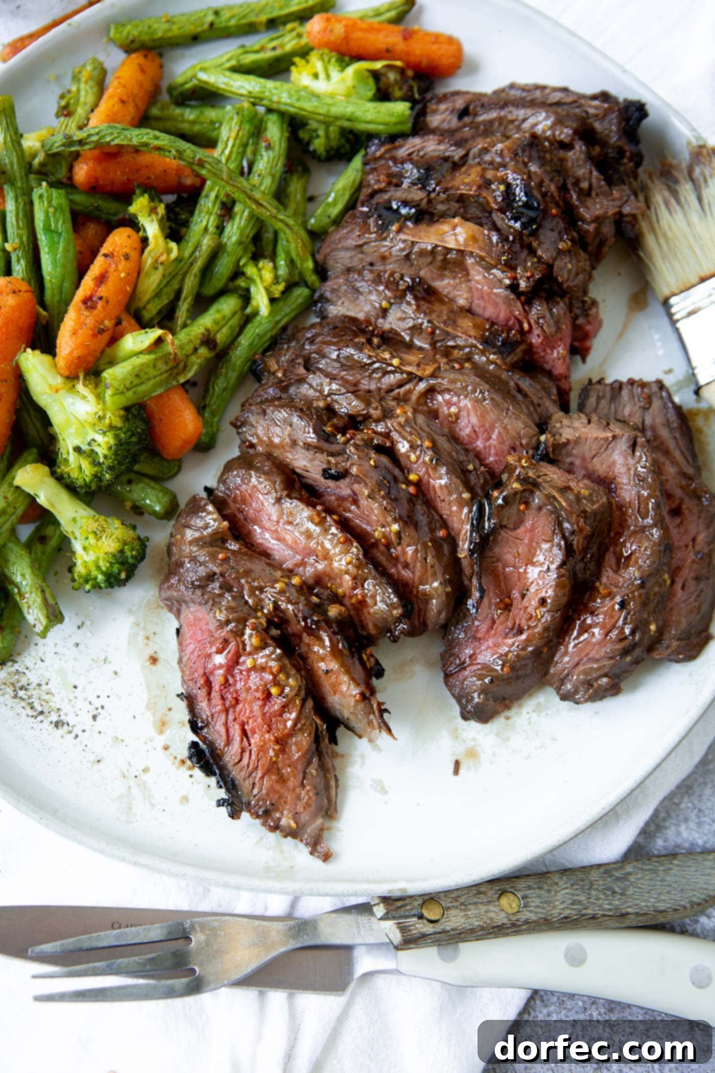 A photo of a sliced medium rare hanger steak on a white plate with roasted baby carrots, broccoli and green beans.