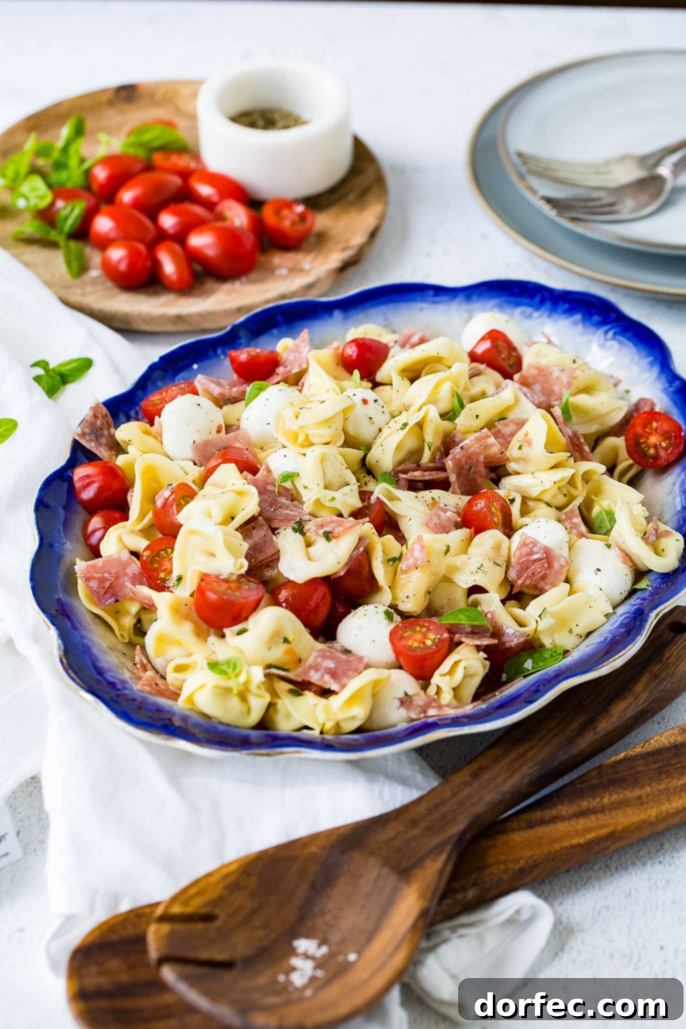 5 Minute Italian Salami Tortellini Salad A blue edged serving dish of 5 minuted Italian salami tortellini salad. There are chunks of salami and slices of tomato in the tortellini. There are also mozzarella balls in the salad. A cutting board with grape tomatoes is in the background.