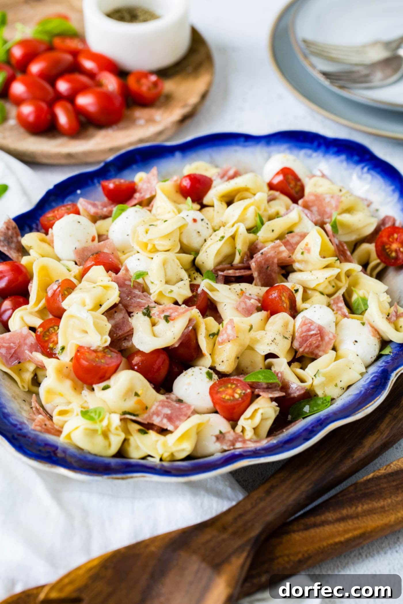 5 Minute Italian Salami Tortellini Salad A blue edged serving dish of 5 minute salami tortellini salad. There are chunks of salami, sliced grape tomatoes, and mozzarella balls mixed in with the tortellini. A wooden cutting board with grape tomatoes is in the background.