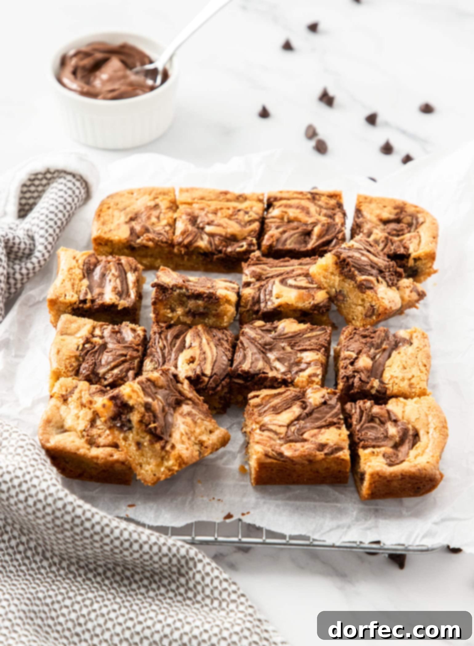 A batch of nutella blondies on a sheet of baking paper with a small bowl of nutella in the background, ready to be enjoyed.