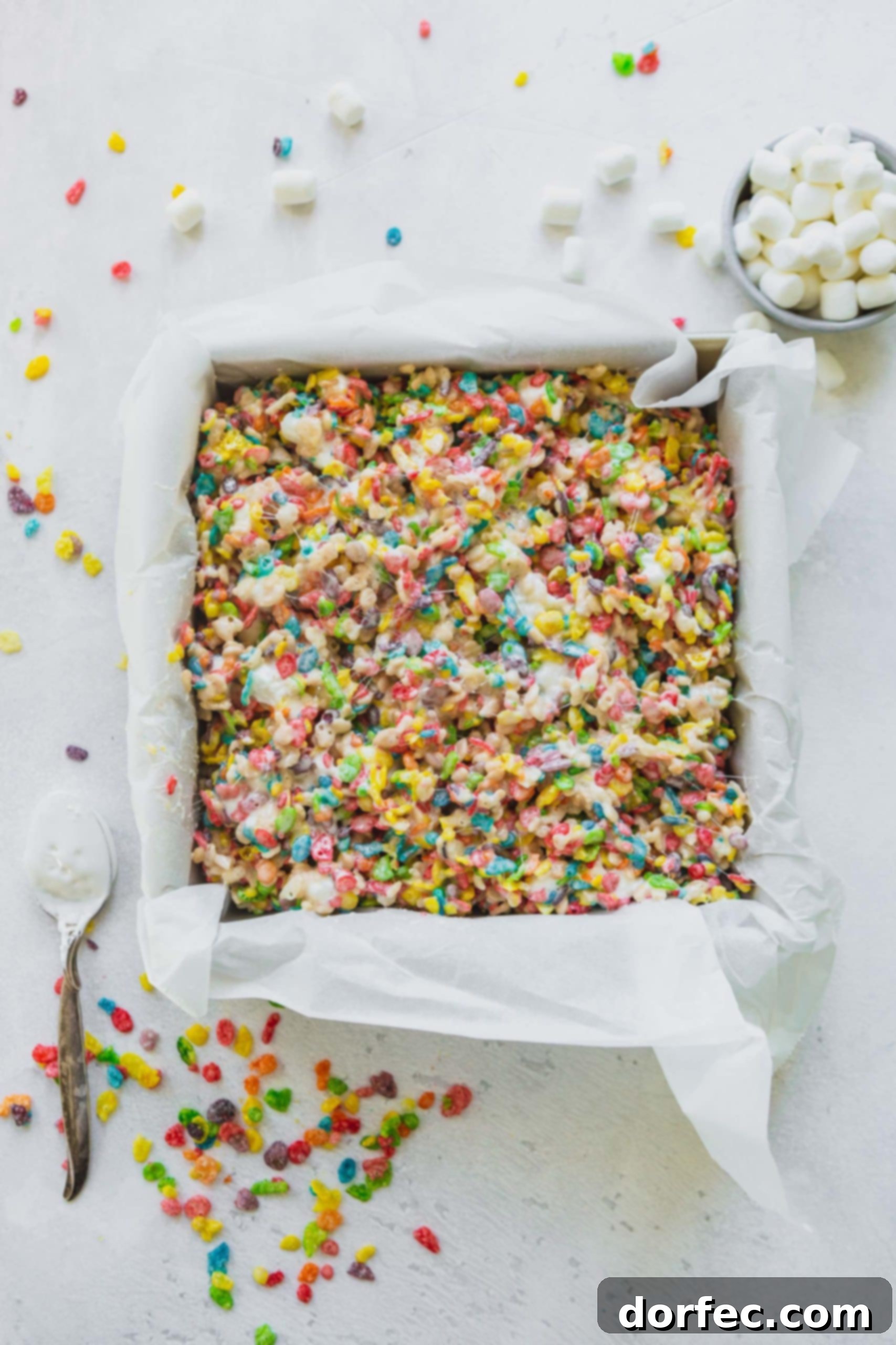 A parchment-lined baking pan filled with freshly made Fruity Pebbles Rice Krispie treats, with colorful cereal pieces scattered around.