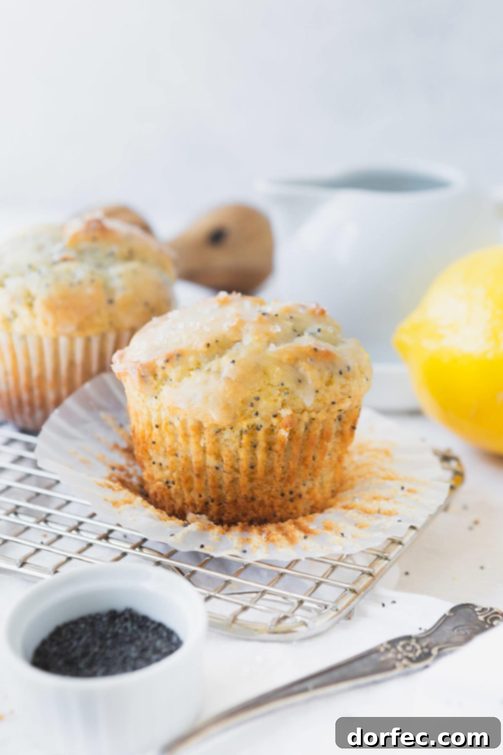 Lemon Poppy Seed Muffins A close-up shot of a lemon poppy seed muffin with its paper liner partially peeled, revealing its soft, golden interior. The muffin is generously covered in a bright lemon glaze. In the blurred background, another muffin, fresh lemons, and a container of poppy seeds complete the inviting scene.