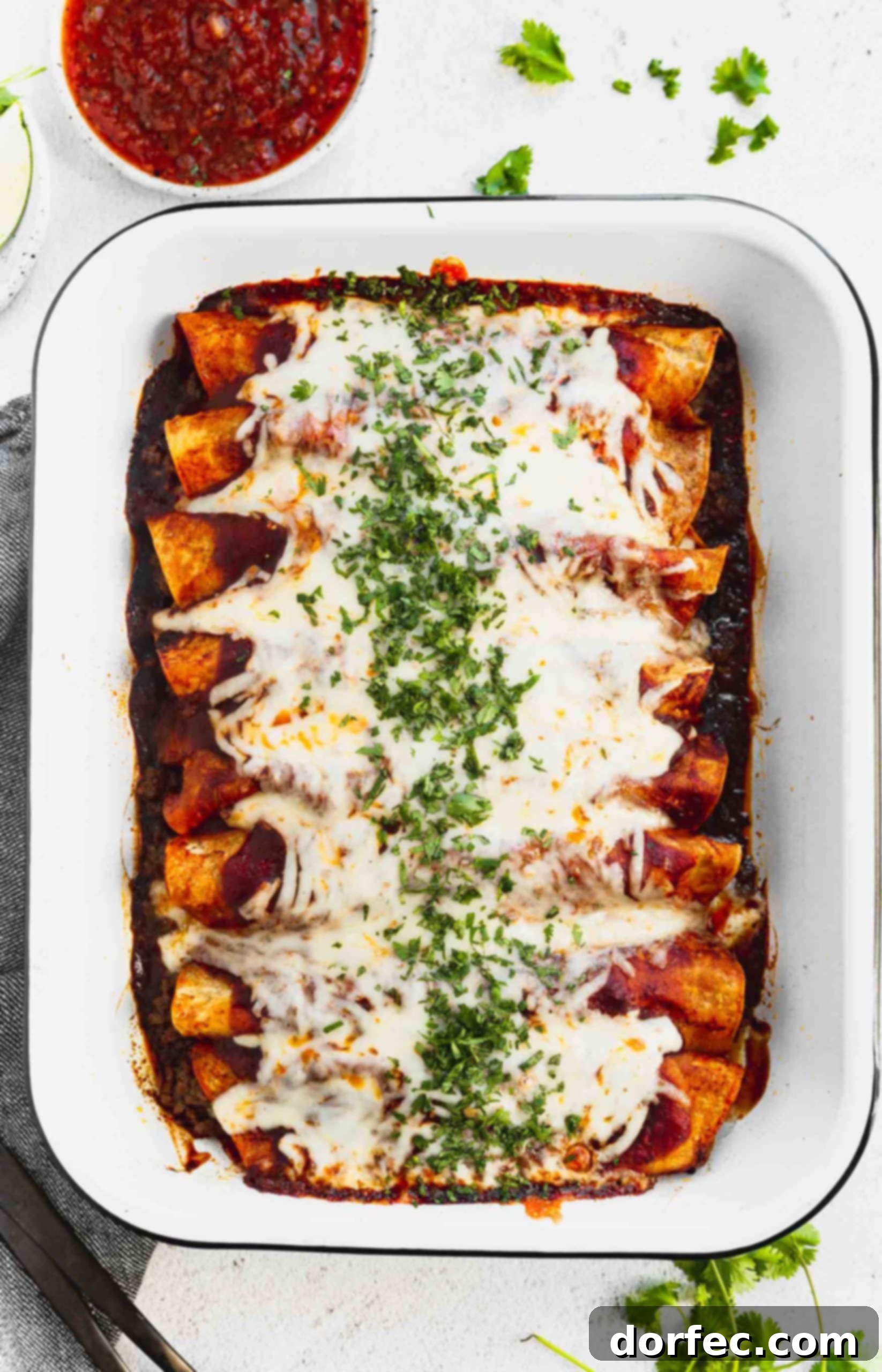 a photo of a pan of ground beef enchiladas from overhead covered in melted cheese and chopped cilantro with a cup of salsa next to the pan.