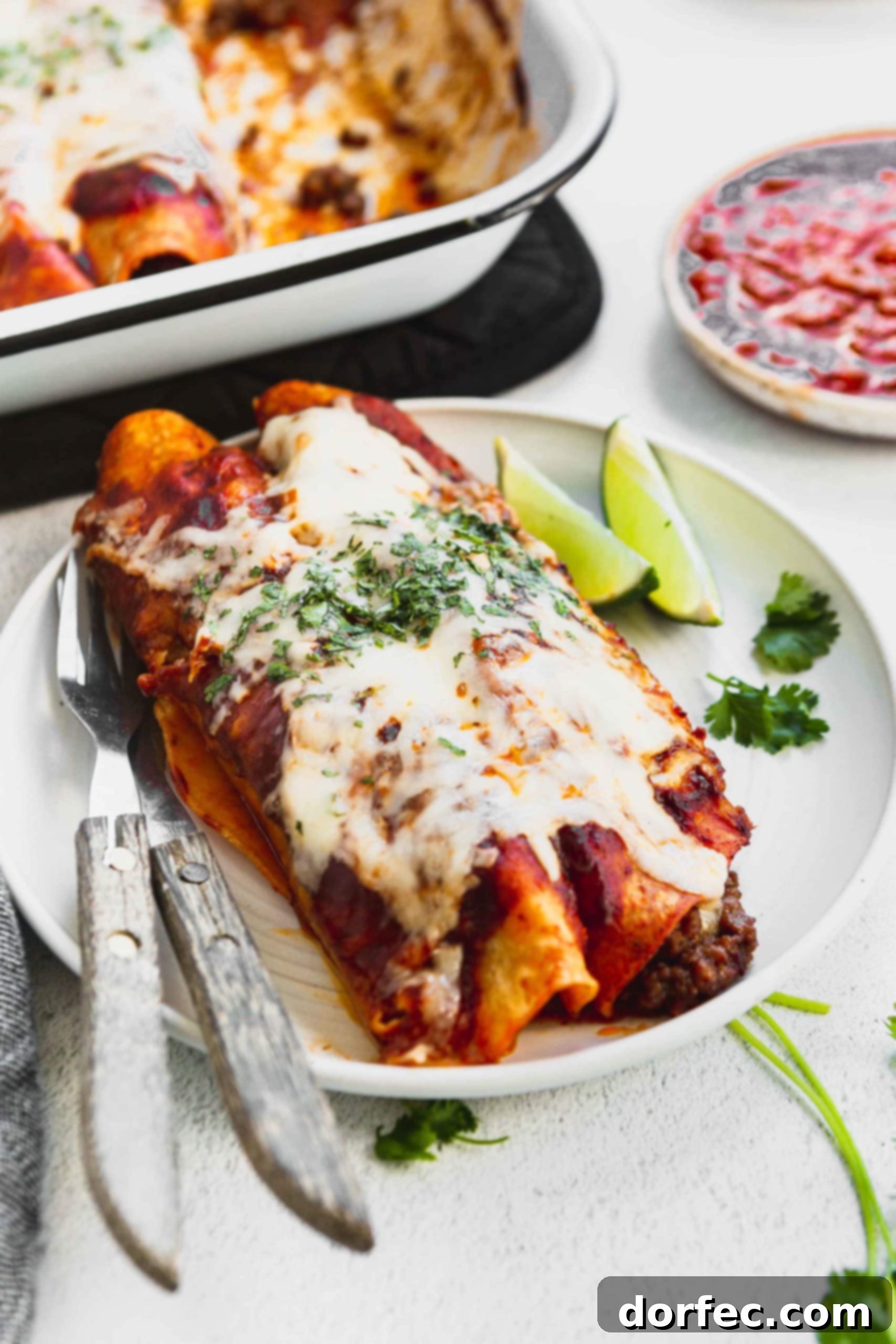 A photo of two ground beef enchiladas on a white plate with two lime wedges on the side.