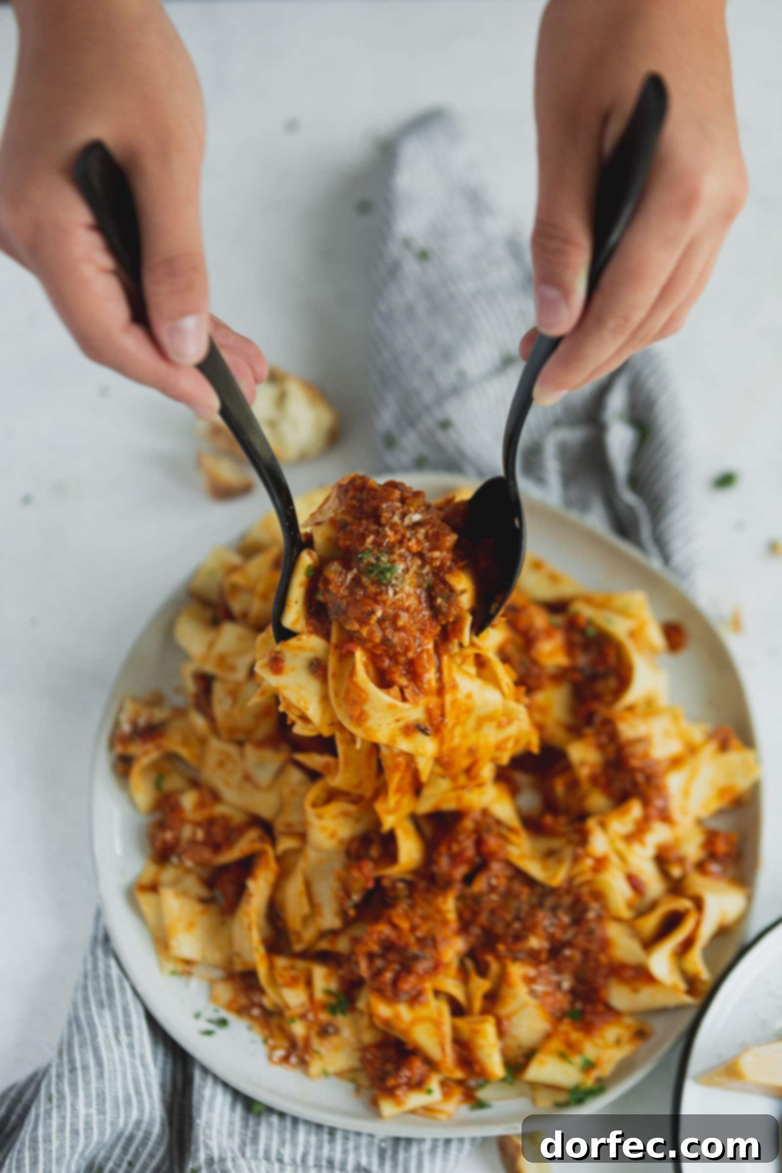 A serving plate piled with pasta that is topped with vegan bolognese sauce. It is being served up with two spoons.