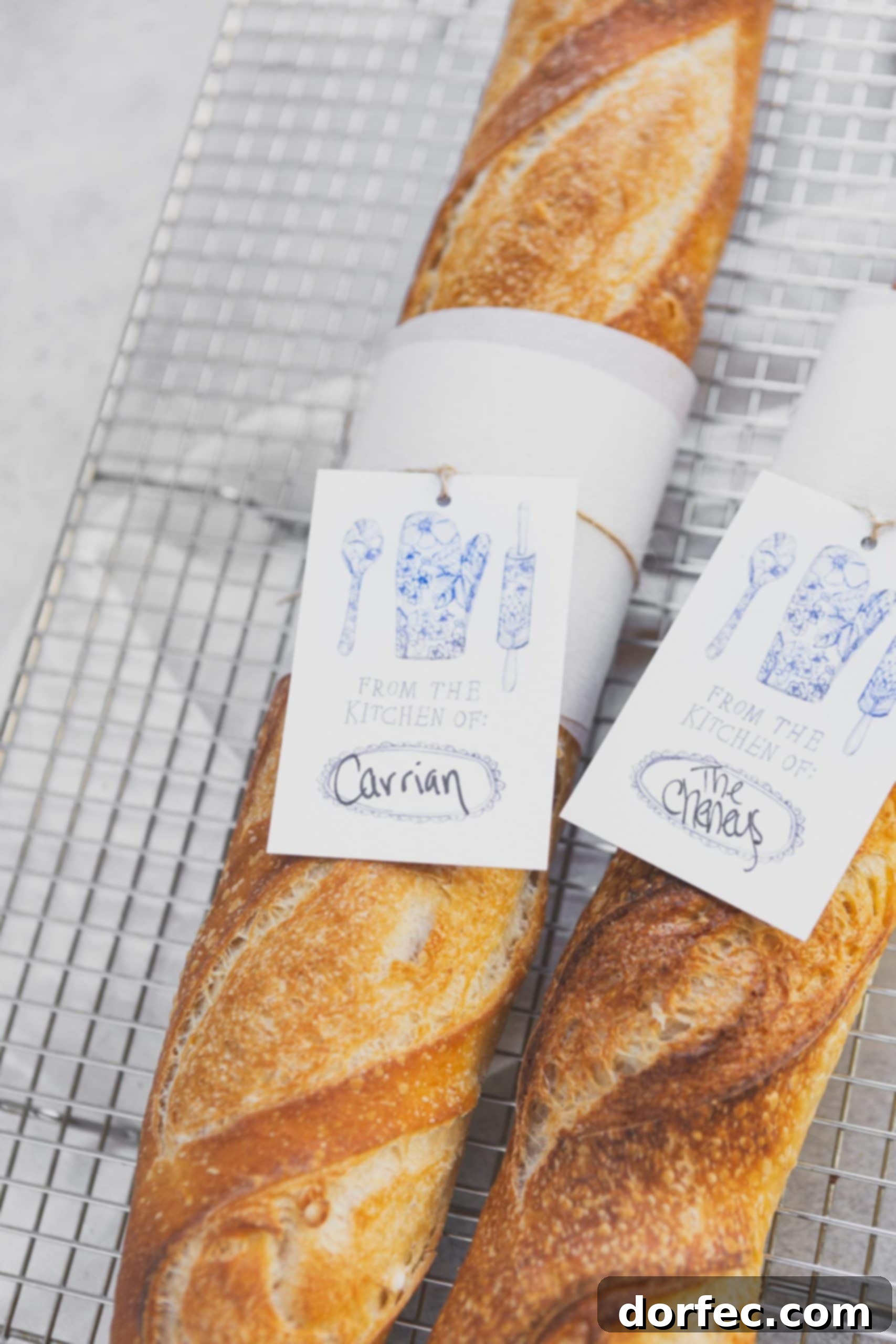 Two loaves of freshly baked baguettes on a wire cooling rack. The baguettes have a paper tag tied around the center.