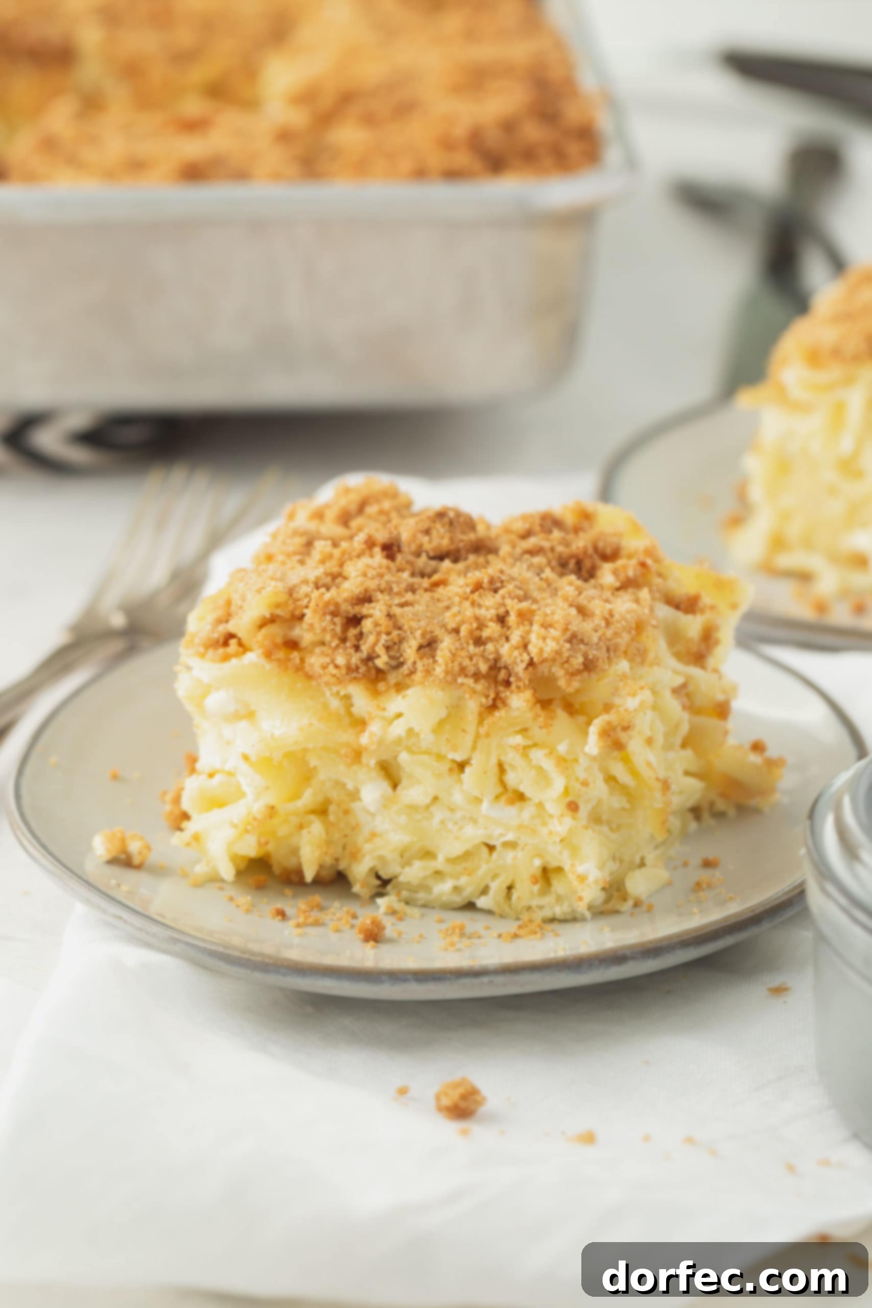 Delicious Jewish Kugel Recipe A beautifully presented serving of Jewish Noodle Kugel on a small white plate, showcasing its golden-brown, creamy noodles, sweet custard, and irresistible graham cracker crumb topping. A larger casserole dish and another serving are blurred in the background.