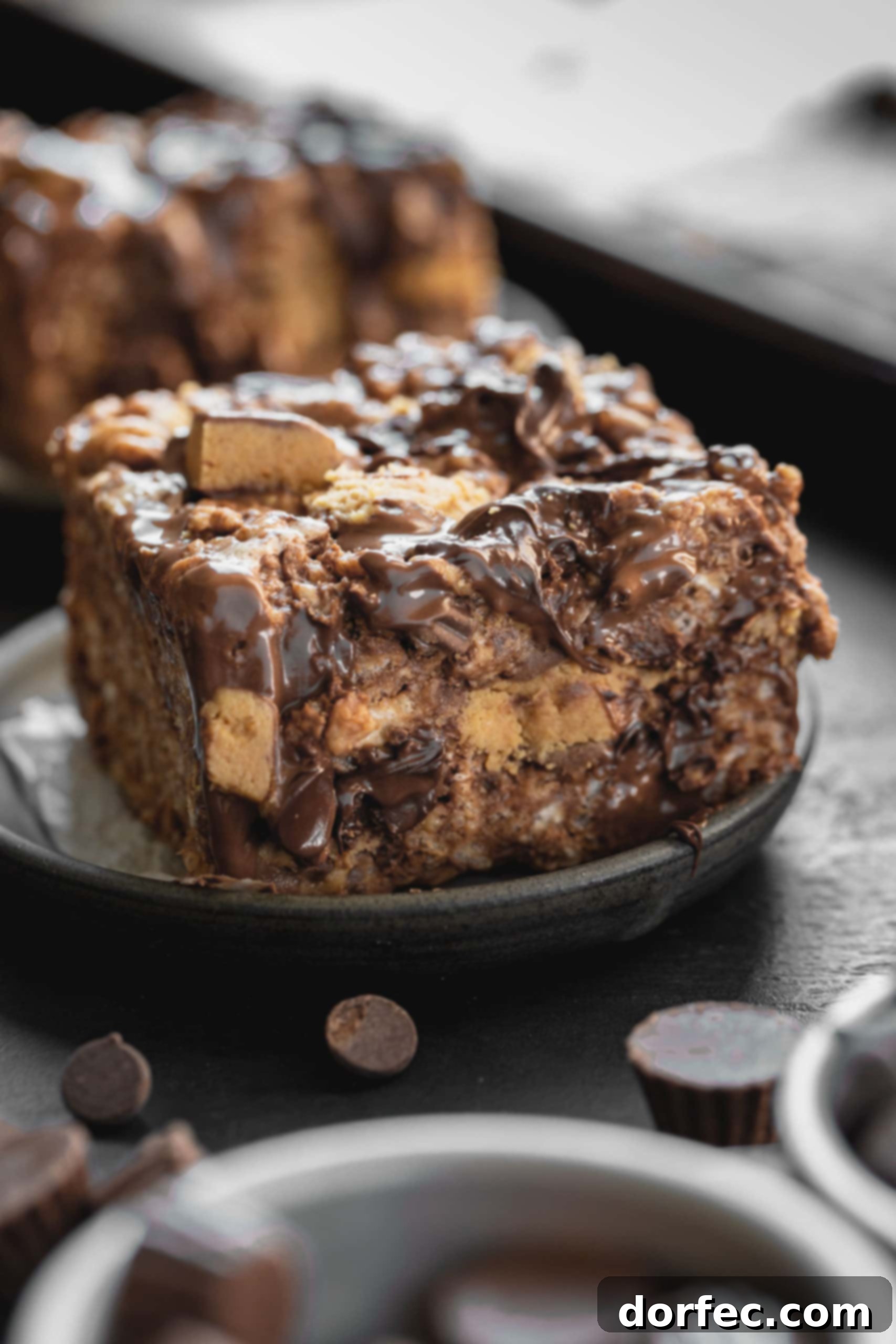 A close-up photo of a single Reese's Rice Krispies Treat on a dark gray plate, loaded with creamy peanut butter, crunchy Rice Krispies, melted chocolate chips, and generous chunks of Reese's Peanut Butter Cups. Several mini Reese's cups are scattered in the foreground.